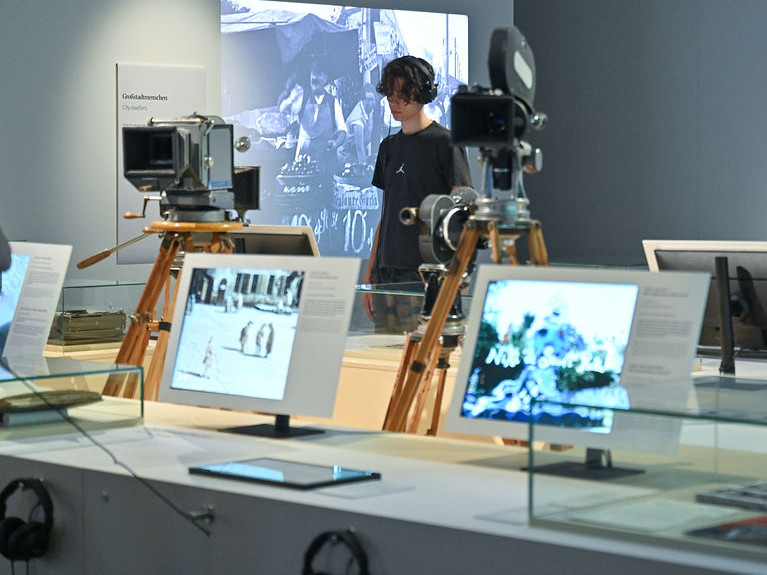 KI generiert: Das Bild zeigt eine Ausstellung mit historischen Kameras und Bildschirmen, auf denen Fotos und Filme präsentiert werden. Eine Person mit Kopfhörern betrachtet die Exponate.