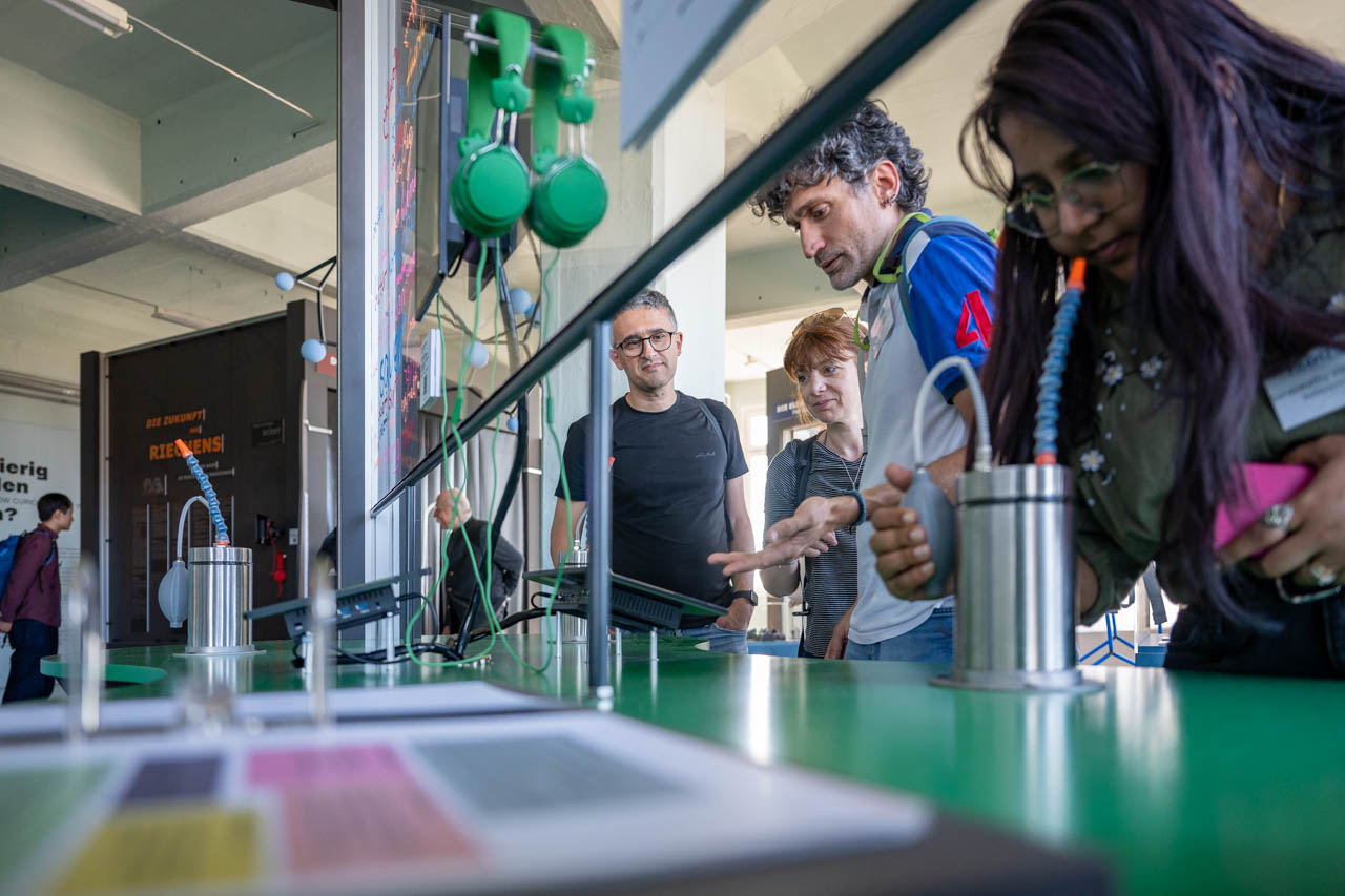 KI generiert: Das Bild zeigt eine Gruppe von Menschen, die sich ein Display mit technischen Geräten in einem modernen Ausstellungsraum ansehen. Sie scheinen interessiert und diskutieren möglicherweise über das Gezeigte.