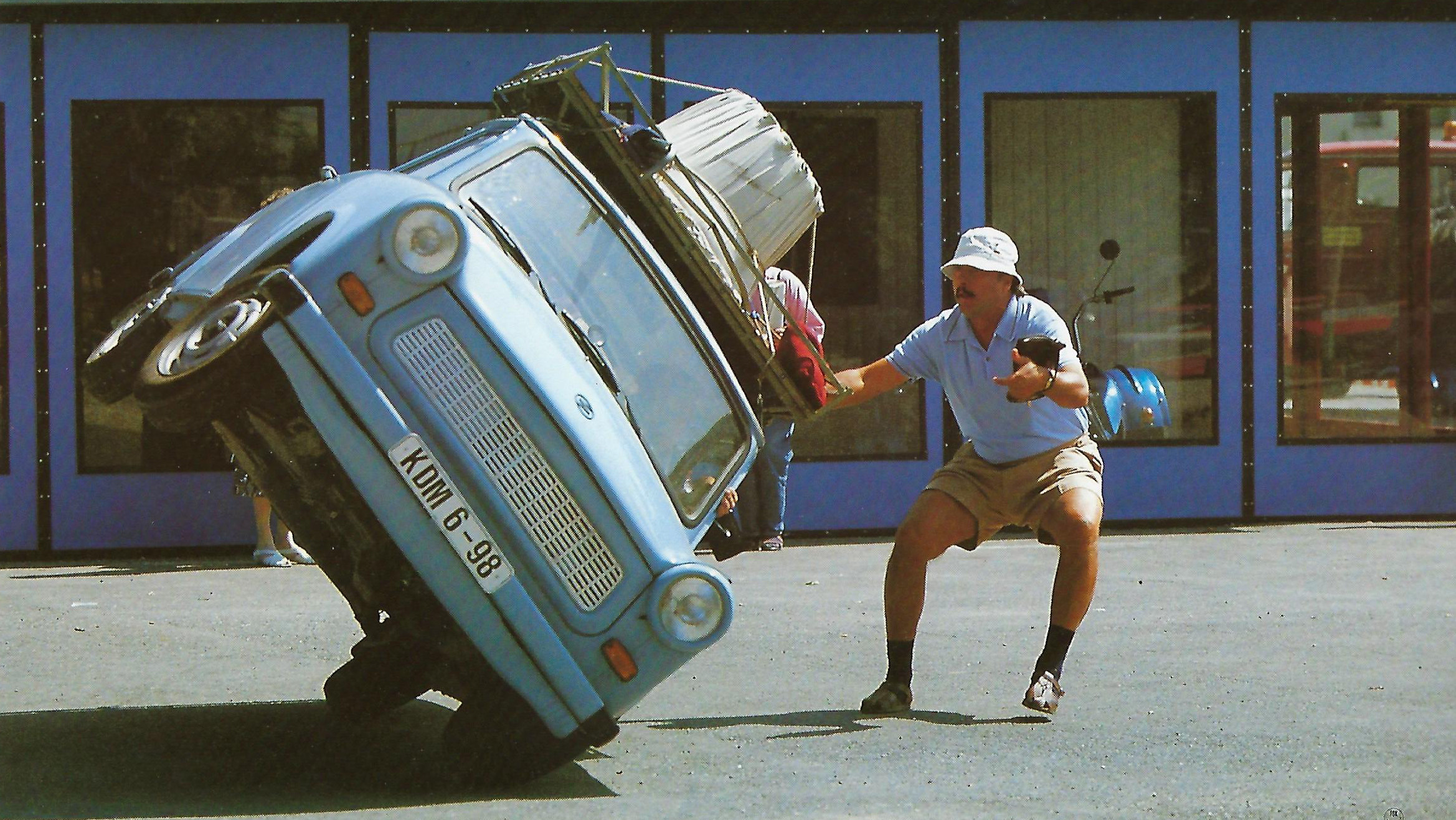 KI generiert: Ein Mann stützt ein blaues Auto, das auf zwei Rädern kippt, während die Aufschrift "Go Trabi Go" im Hintergrund zu sehen ist. Der Hauptinhalt des Bildes ist die humorvolle und dynamische Darstellung eines einradigen Fahrmanövers.