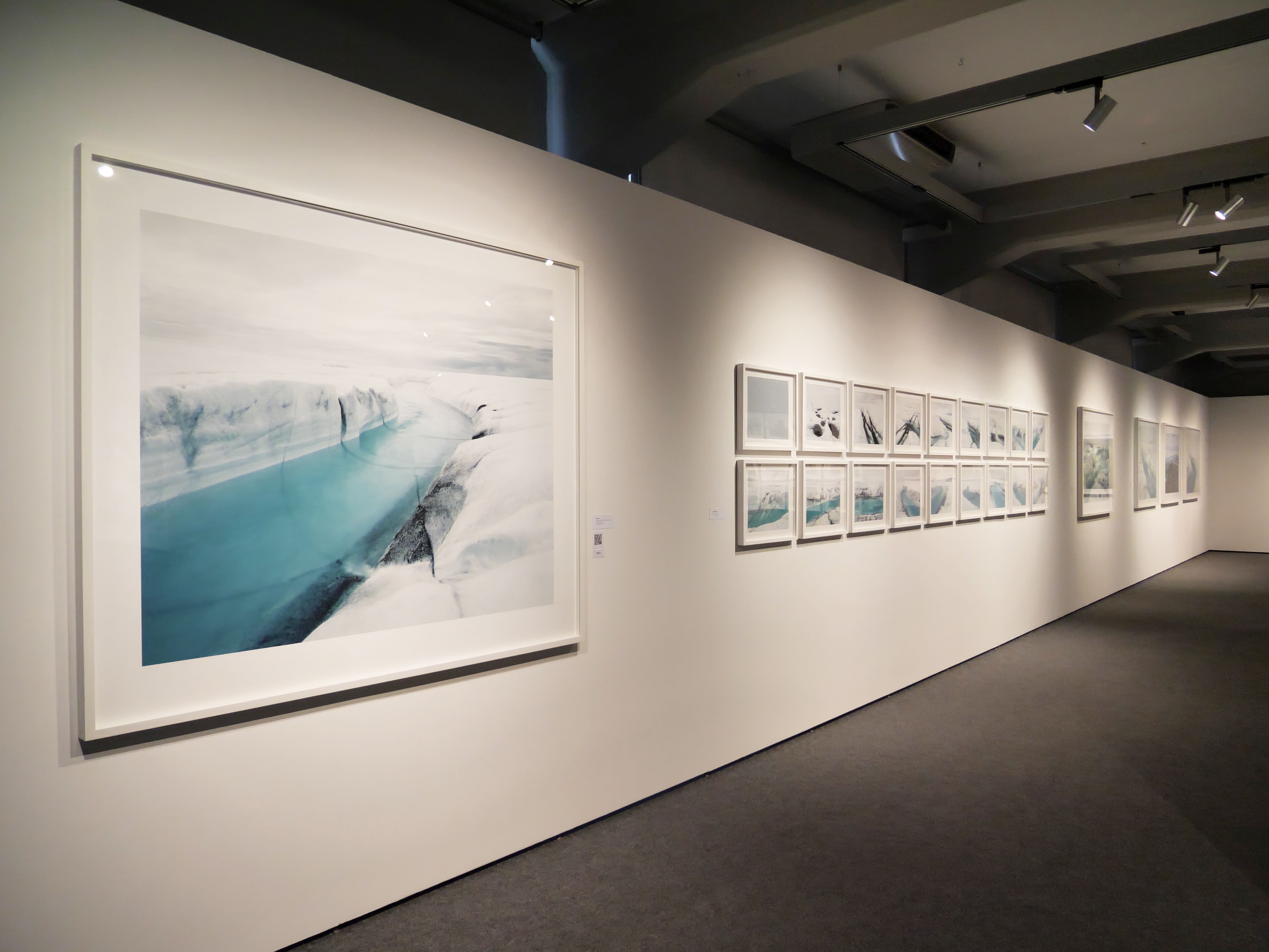 KI generiert: Das Bild zeigt eine Kunstgalerie mit großen Fotoprints an der Wand, die hauptsächlich Landschaften und Naturaufnahmen darstellen. Der Hauptinhalt des Bildes ist eine große Fotografie eines eisigen Geländes mit blauem Wasser, die auf der linken Seite prominent ausgestellt ist.