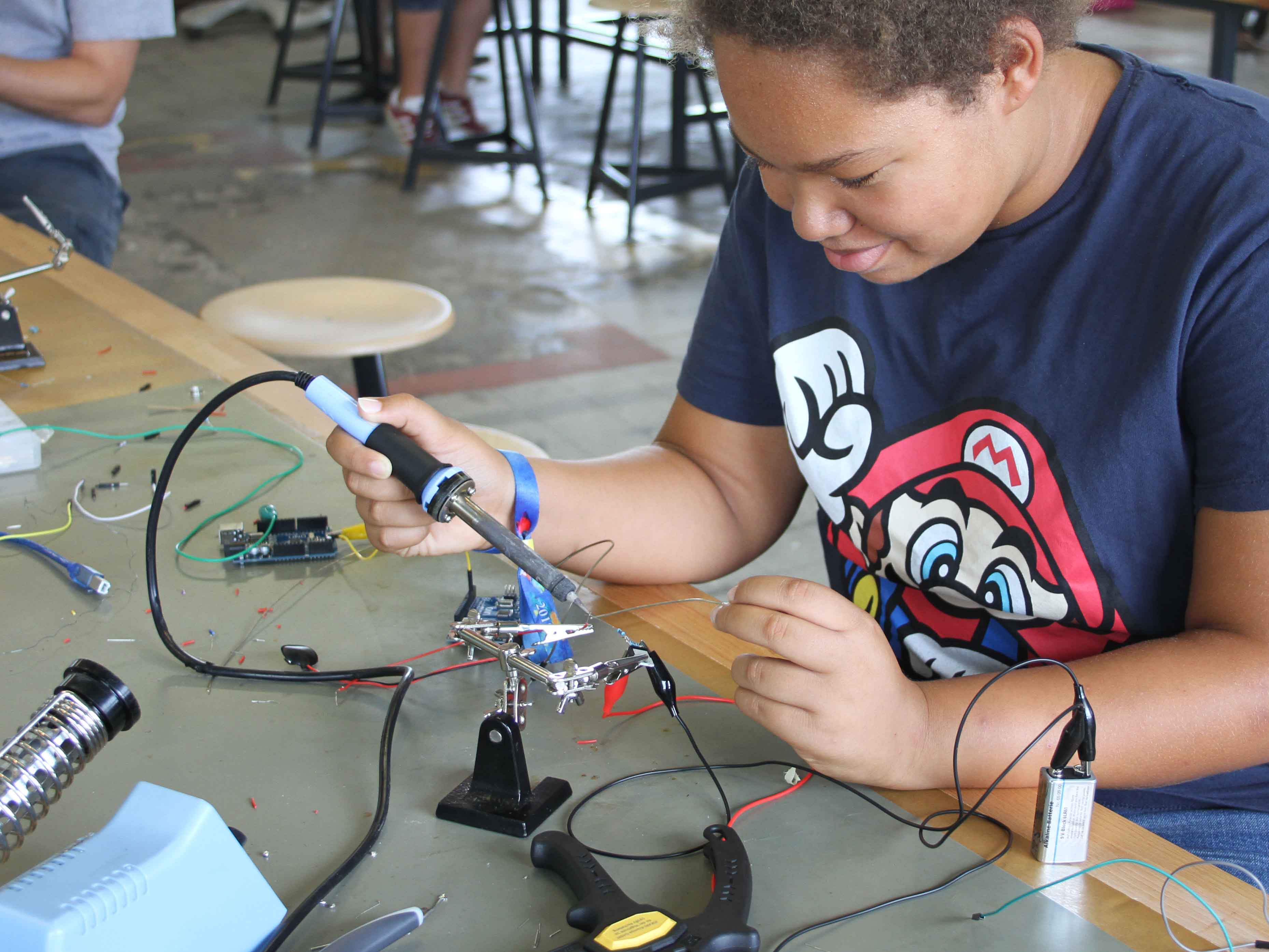KI generiert: Das Bild zeigt eine Person, die konzentriert an elektronischen Komponenten arbeitet, vermutlich beim Löten eines Schaltkreises. Umgeben von Werkzeugen und Kabeln sitzt sie an einem Tisch in einem Workshop-ähnlichen Raum.