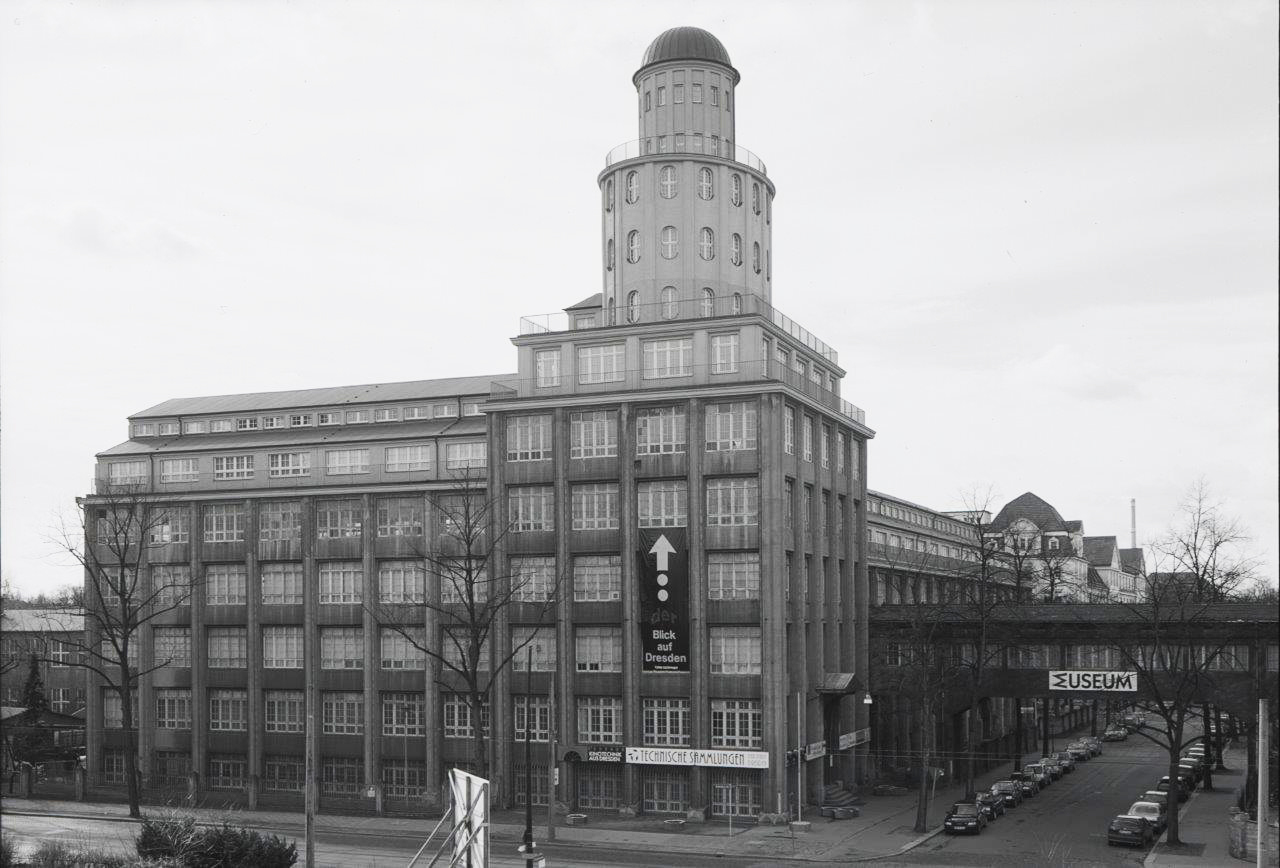 KI generiert: Das Bild zeigt ein großes, mehrstöckiges Gebäude mit einem markanten runden Turm. Am unteren Rand des Gebäudes befinden sich Schilder, die auf ein "Museum" und "Technische Sammlungen" hinweisen.