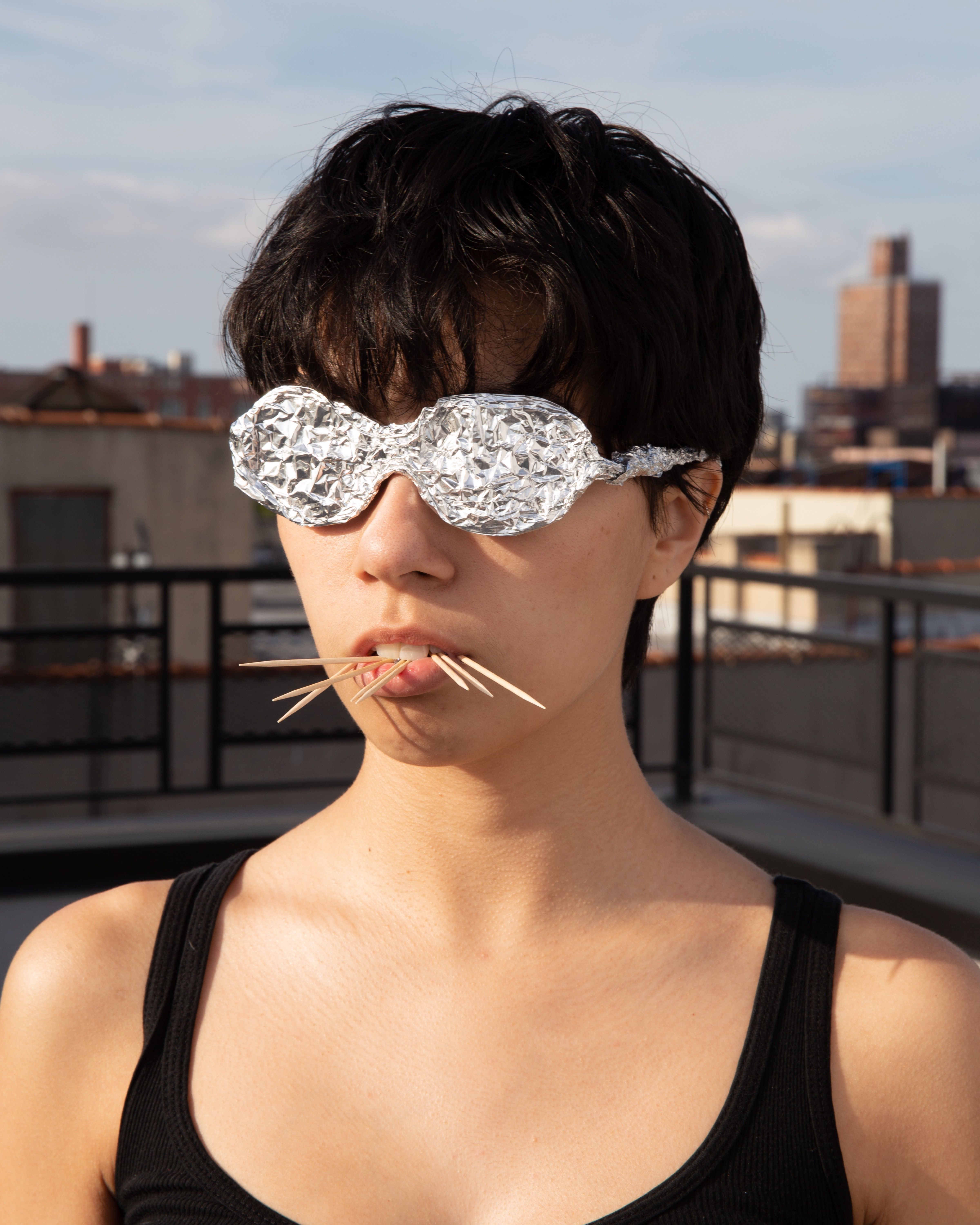 KI generiert: Das Bild zeigt eine Person, die eine Brille aus Aluminiumfolie trägt und mehrere Zahnstocher im Mund hat. Der Hintergrund deutet auf eine städtische Umgebung hin.