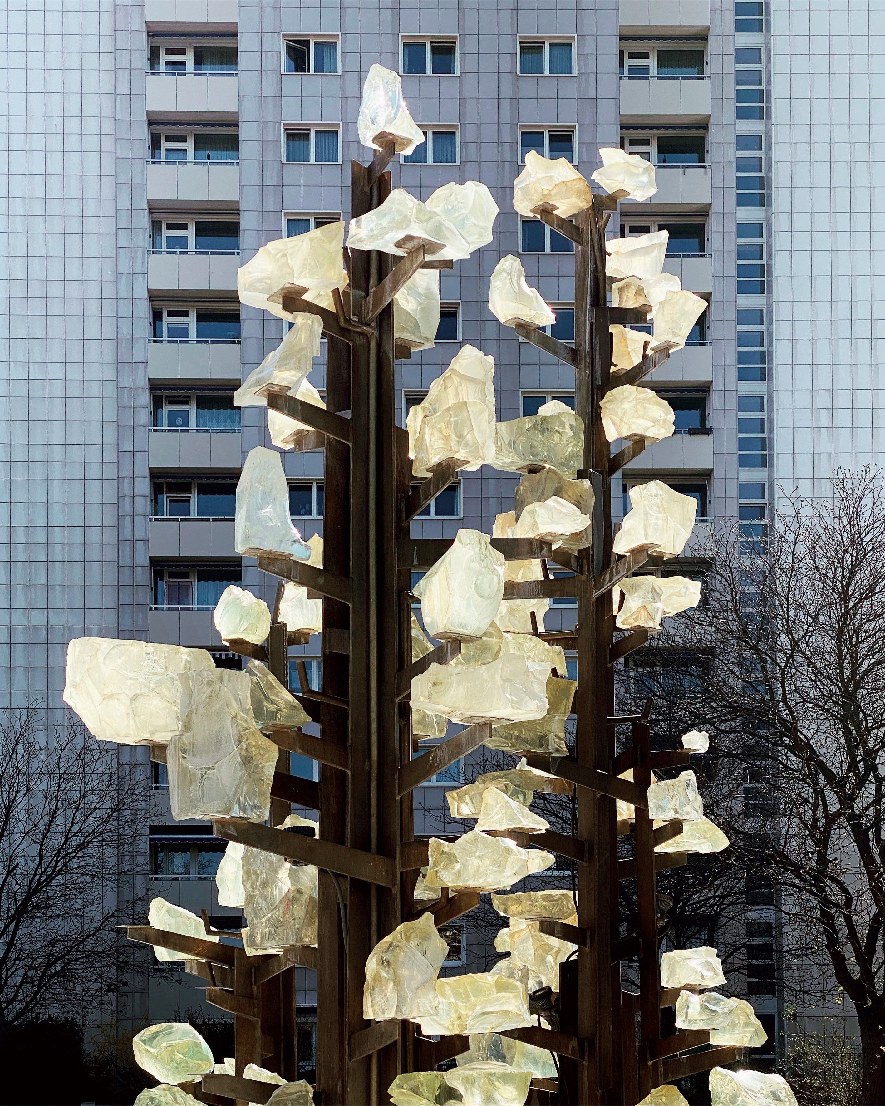 KI generiert: Das Bild zeigt eine moderne Skulptur mit verzweigten Strukturen und leuchtenden, gläsernen Elementen im Vordergrund, während im Hintergrund ein hohes Wohngebäude sichtbar ist. Die Skulptur steht vermutlich im Freien, umgeben von Bäumen.