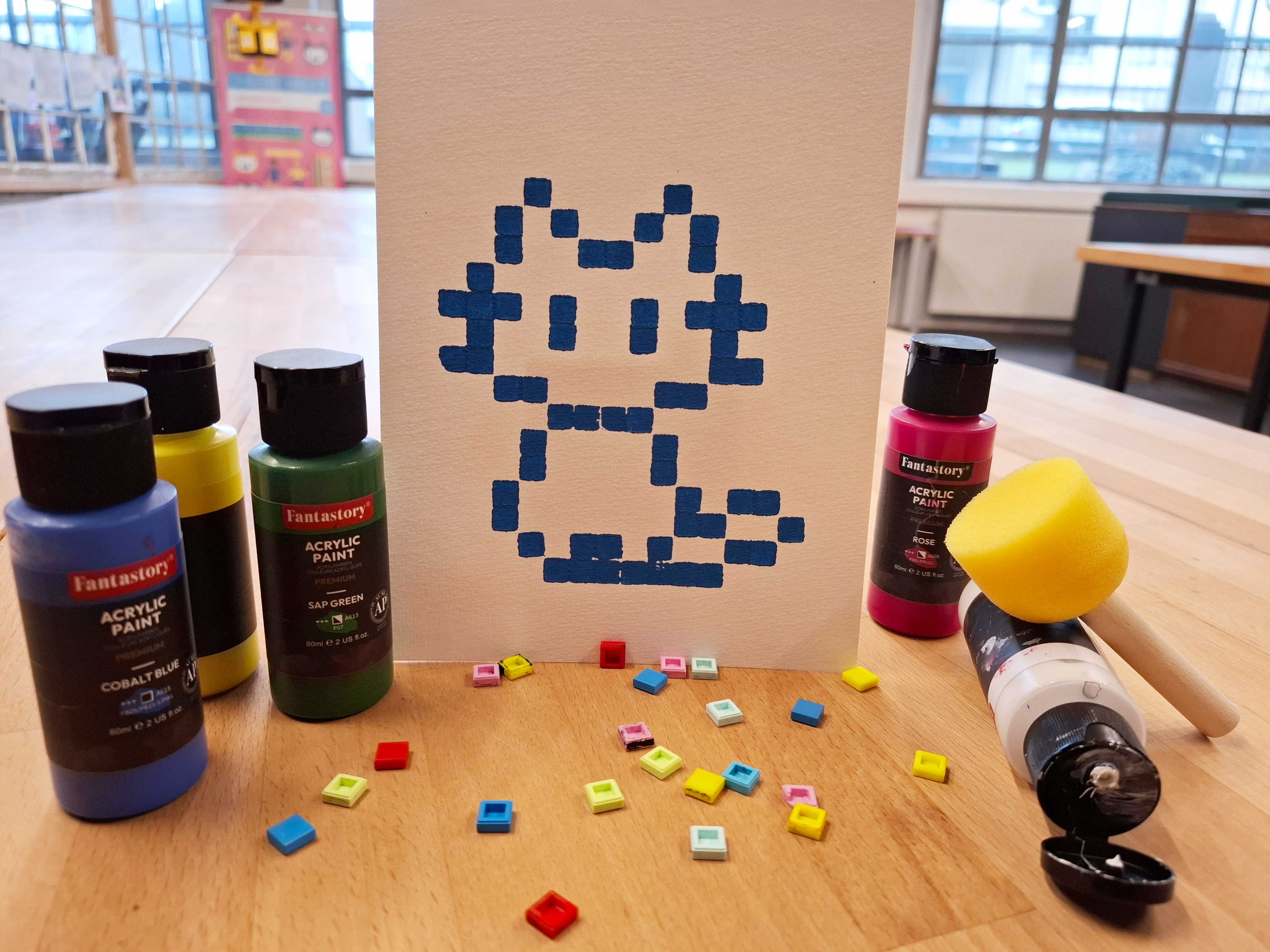 AI generated: The image shows a canvas painting with a pixel-like, blue cat figure, surrounded by acrylic paints and colourful mosaic tiles on a wooden table. The main content of the image is the creative depiction of this digital cat.