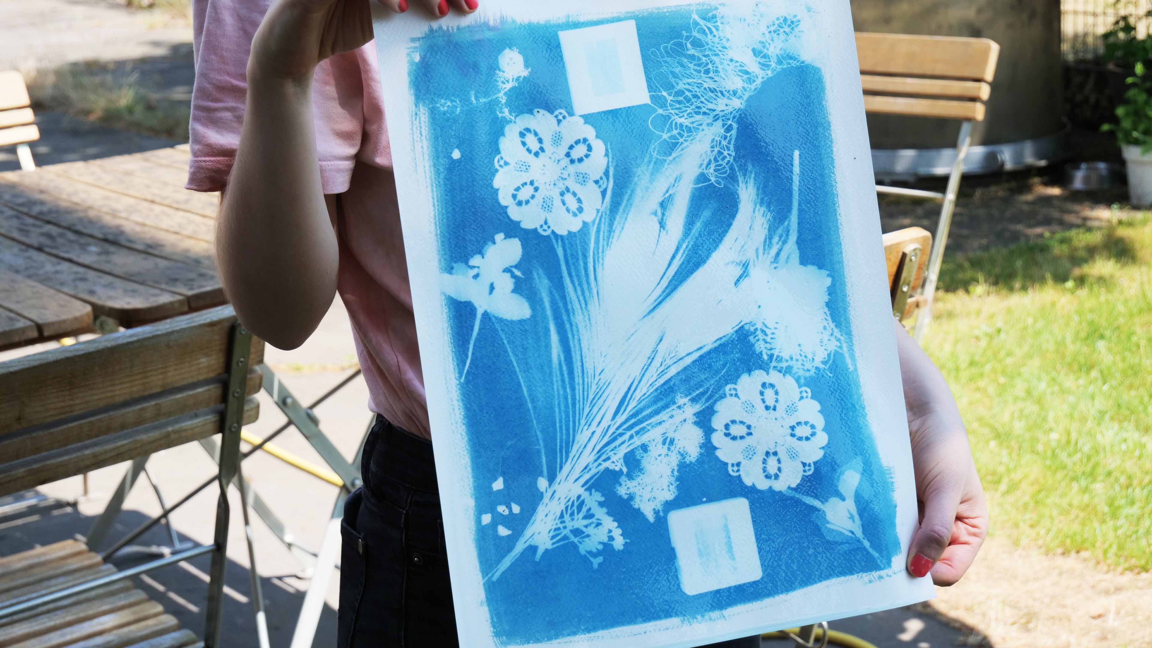 KI generiert: Eine Person hält ein Cyanotypie-Bild hoch, das weiße Blumen- und Pflanzenmuster auf einer blauen Fläche zeigt. Im Hintergrund sind Gartenmöbel erkennbar.