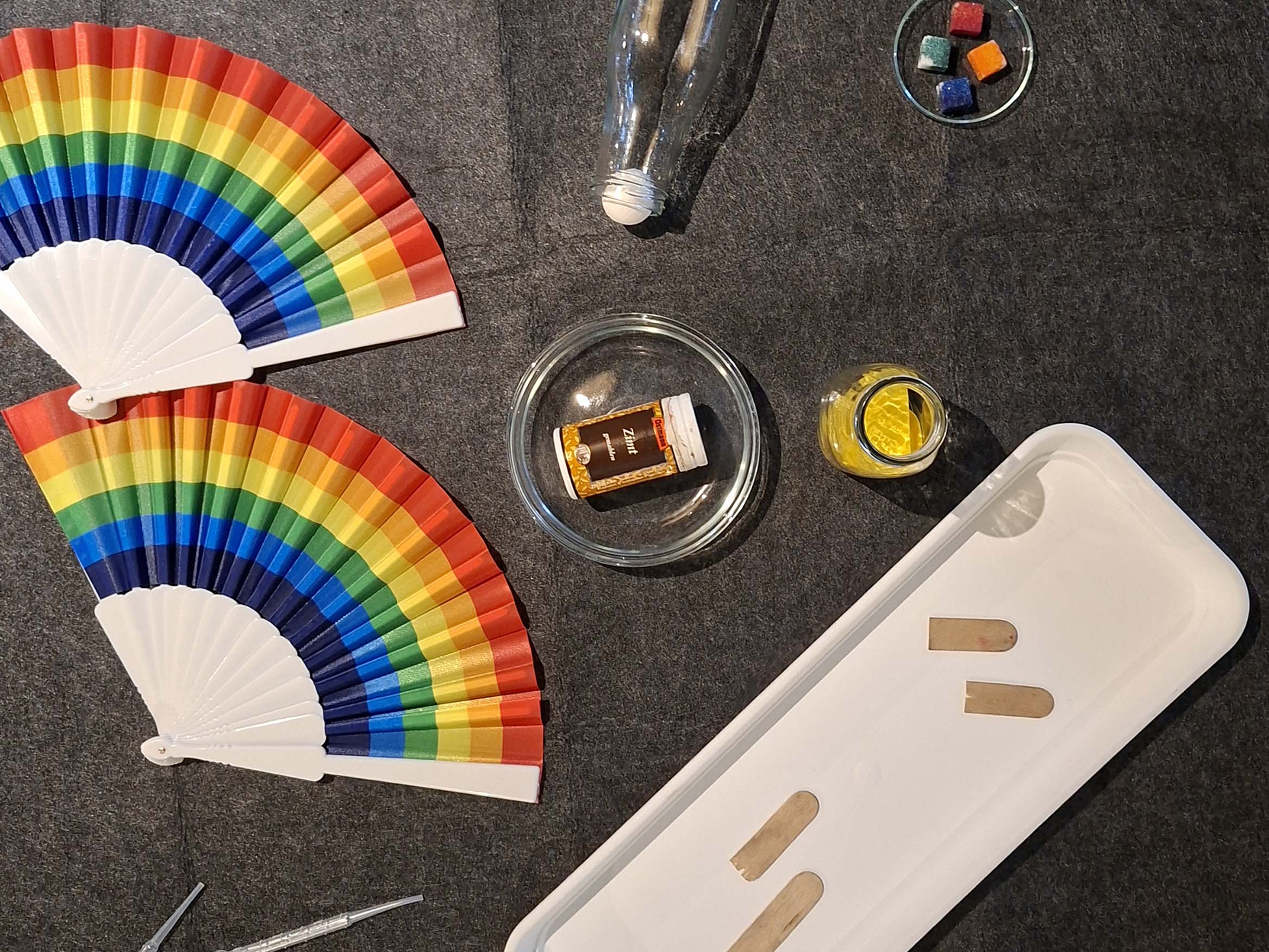 KI generiert: Das Bild zeigt eine Anordnung von verschiedenen Gegenständen auf einer dunklen Oberfläche, darunter zwei Fächer mit Regenbogenfarben, ein Glasgefäß mit einer Flasche darin und bunte Würfel in einer Schale. Es handelt sich um eine künstlerische Komposition mit Alltagsgegenständen.