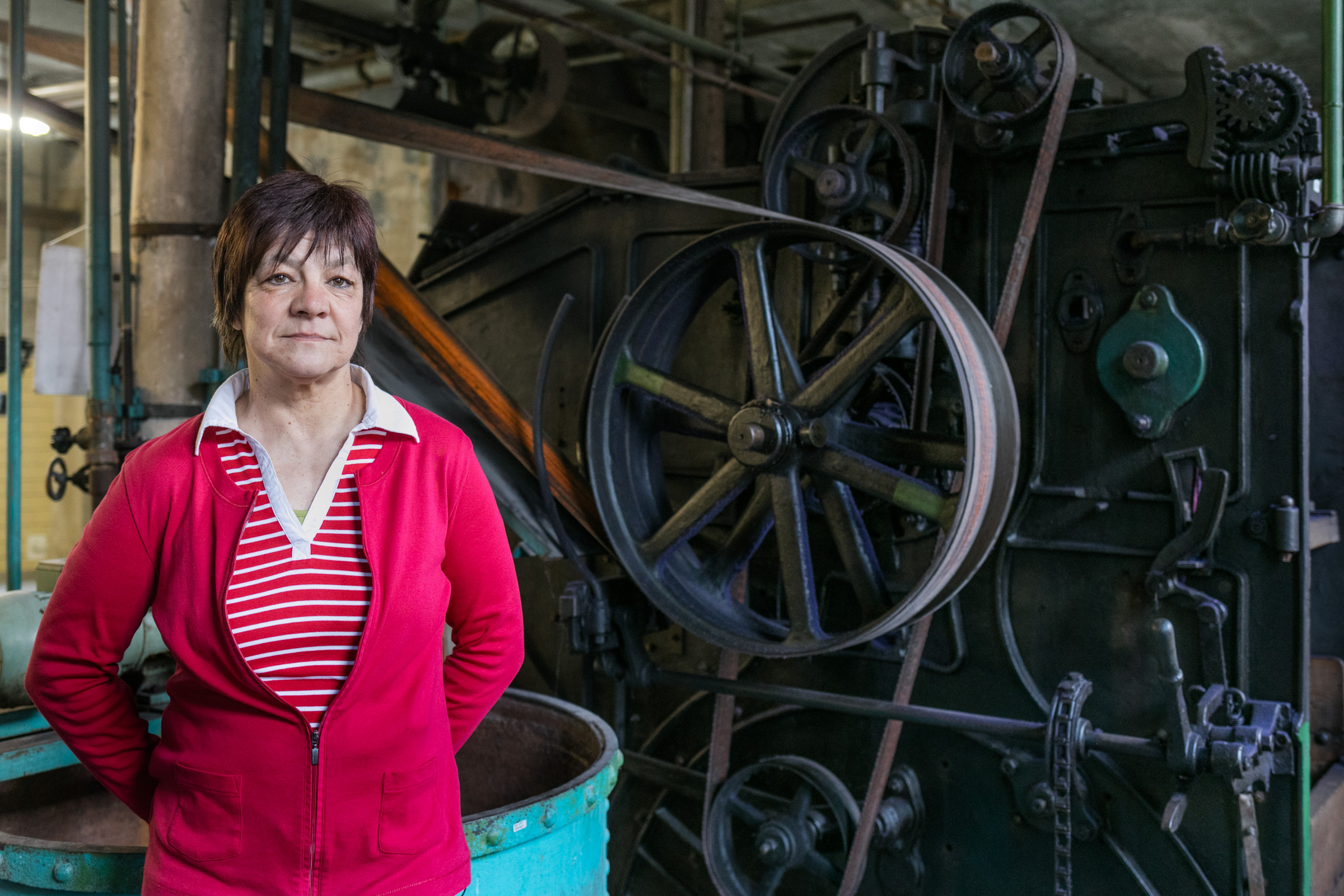 KI generiert: Das Bild zeigt eine Frau in einem roten Pullover, die vor einer großen, alten Industriemaschine steht. Der Hintergrund ist eine industrielle Umgebung mit verschiedenen mechanischen Komponenten und Riemenantrieben.