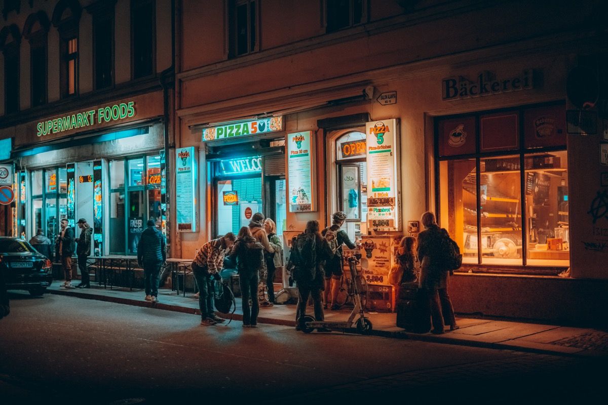 KI generiert: Das Bild zeigt eine abendliche Straßenszene mit Menschen, die sich vor verschiedenen Geschäften aufhalten, darunter ein Supermarkt, eine Pizzeria und eine Bäckerei. Die warmen Lichter der Geschäfte erzeugen eine gemütliche Atmosphäre.