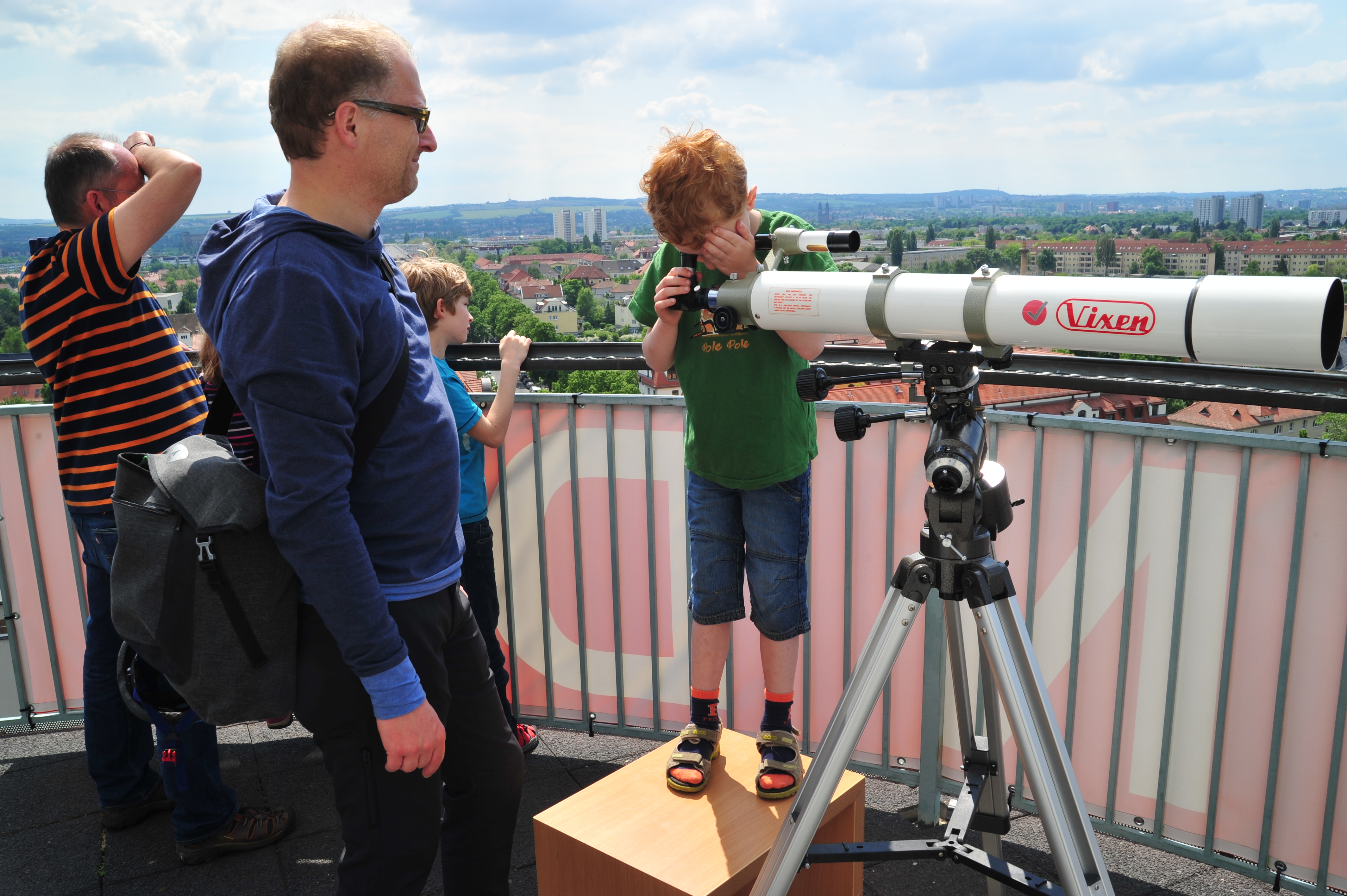 KI generiert: Ein Kind schaut durch ein großes Teleskop, während ein Erwachsener und weitere Kinder in der Nähe zuschauen. Die Szene spielt sich auf einer Aussichtsplattform mit Aussicht auf eine Stadtlandschaft ab.