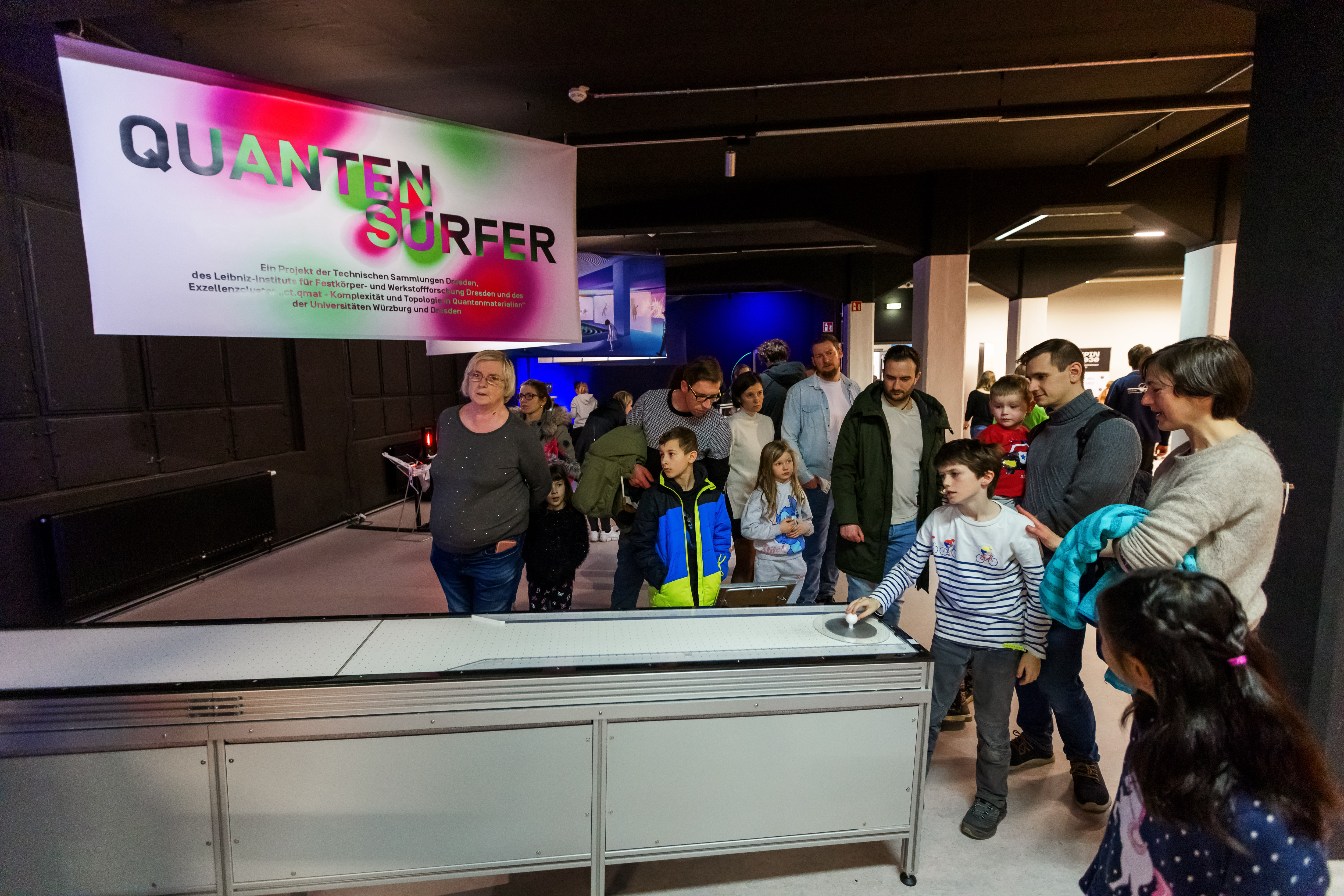 KI generiert: Das Bild zeigt eine Gruppe von Menschen, darunter viele Kinder, die sich in einem Ausstellungsraum versammelt haben, um eine interaktive Vorführung mit dem Titel "Quantensurfer" zu beobachten. Es scheint Teil einer Ausstellung über Technologie oder Wissenschaft zu sein.