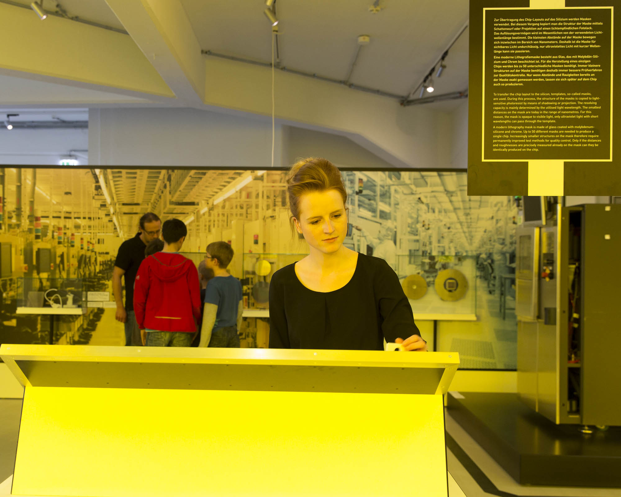 KI generiert: Das Bild zeigt eine Frau, die vor einem großen, beleuchteten Lesepult in einem Technik-Museum oder Ausstellung steht. Im Hintergrund sind einige Besucher sowie technische Exponate zu sehen.