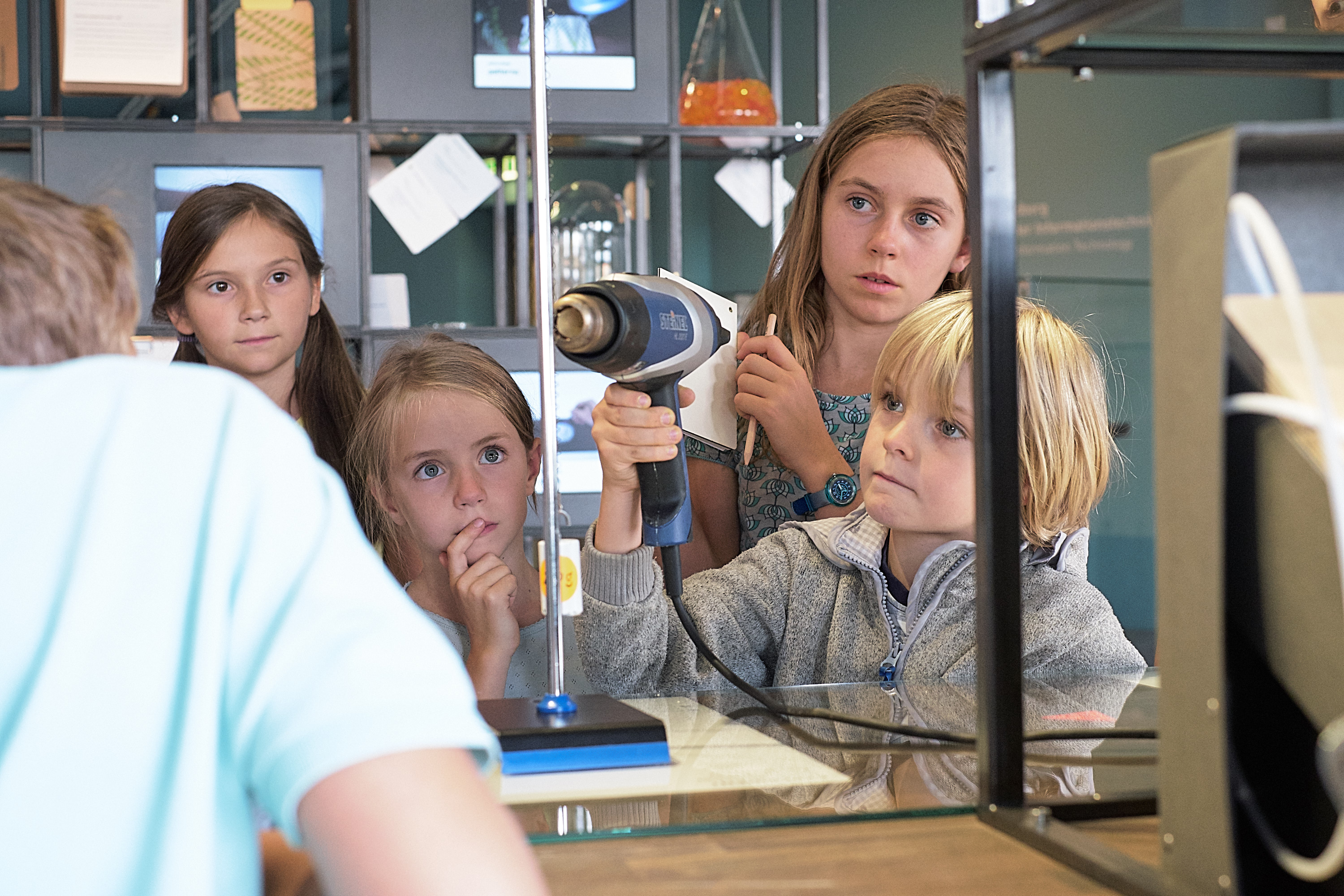 KI generiert: Das Bild zeigt eine Gruppe von Kindern, die in einem wissenschaftlichen Umfeld an einem Experiment oder einer Vorführung teilnehmen. Ein Junge hält ein technisches Gerät, während die anderen Kinder aufmerksam zuschauen.