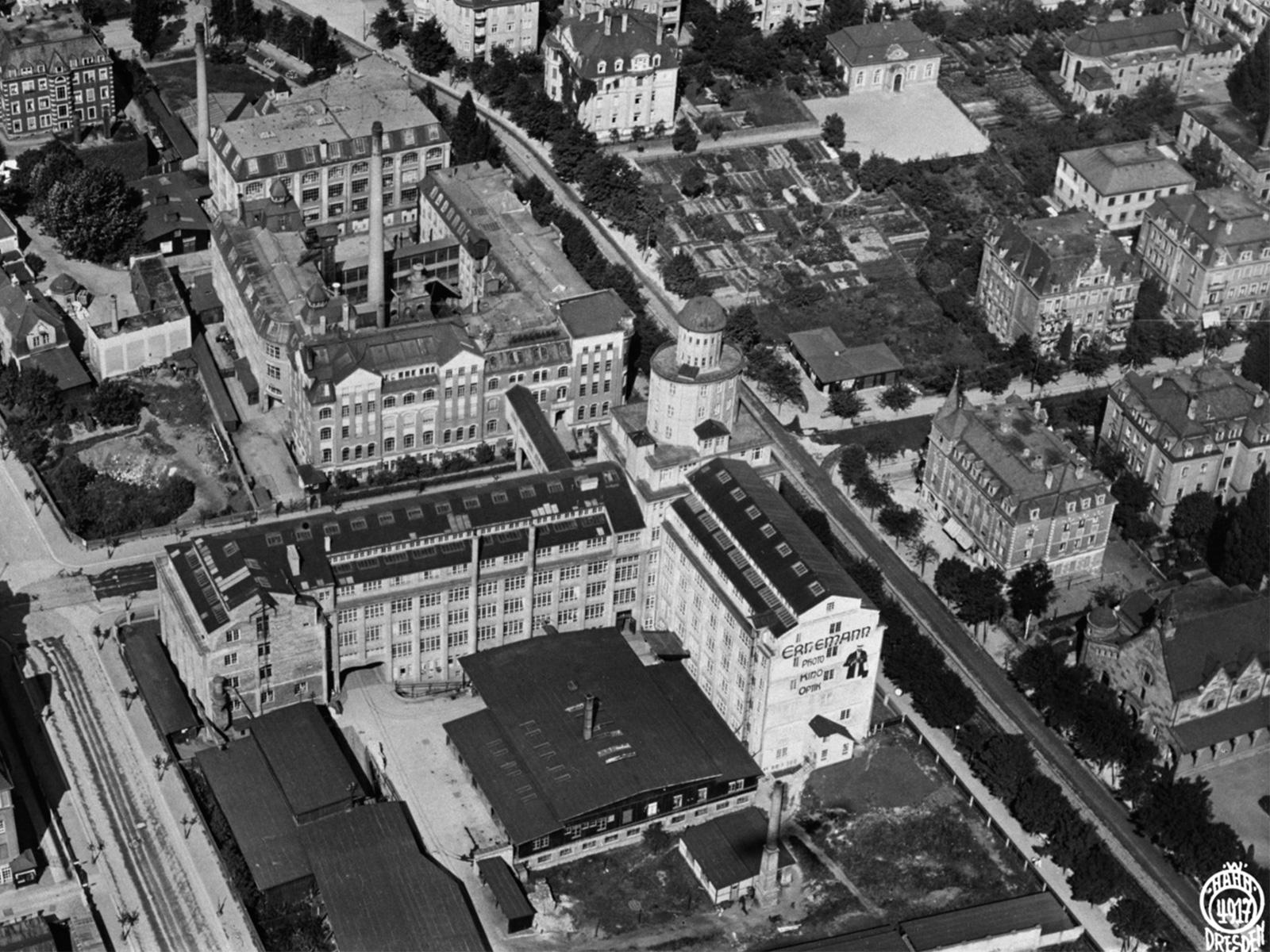 KI generiert: Das Bild zeigt eine Luftaufnahme eines Stadtviertels mit mehreren großen Gebäudekomplexen, darunter ein markantes Gebäude mit Türmen und Schornsteinen. Es handelt sich vermutlich um ein altes Industrie- oder Fabrikgelände, das von Wohnhäusern und Gärten umgeben ist.