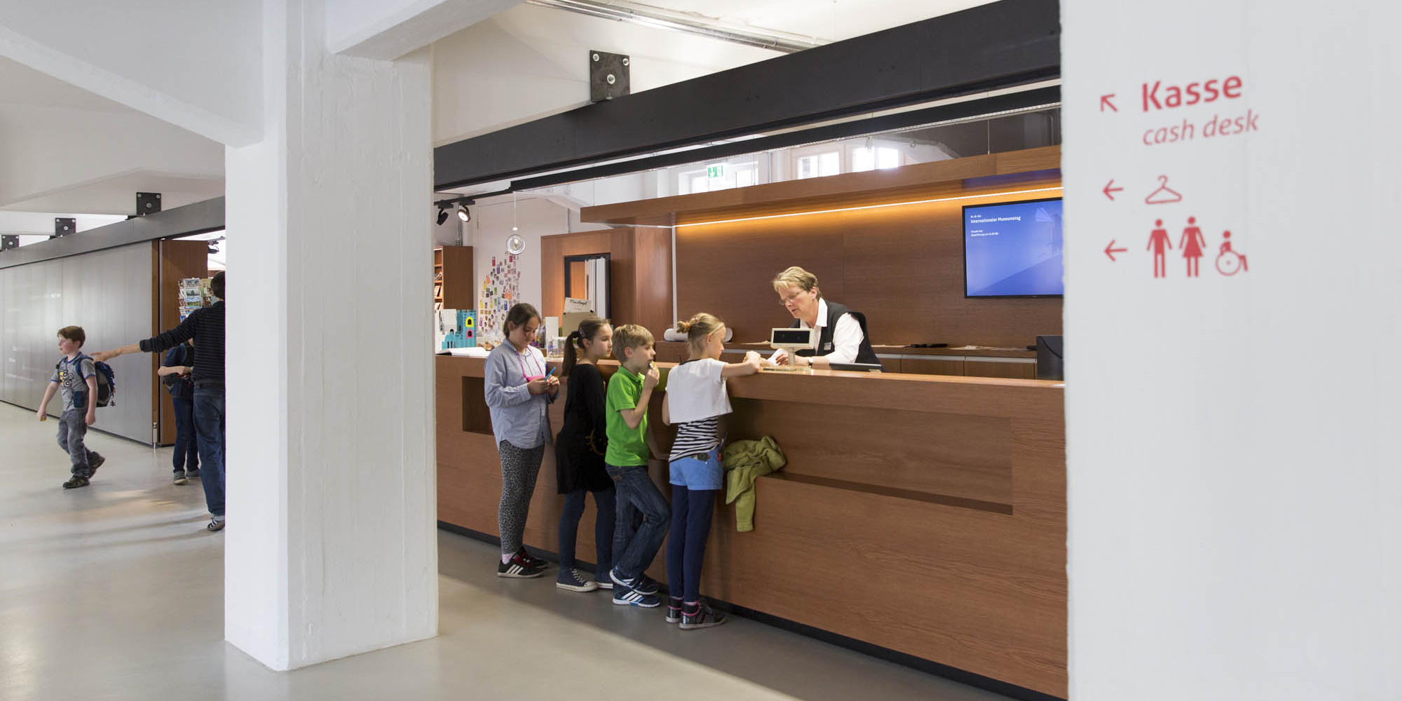 KI generiert: Das Bild zeigt eine Gruppe von Kindern, die an einem Schalter in einer Halle oder einem Foyer von einem Erwachsenen bedient werden. Auf der rechten Seite des Bildes weist ein Schild den Weg zur Kasse und zu den Toiletten.