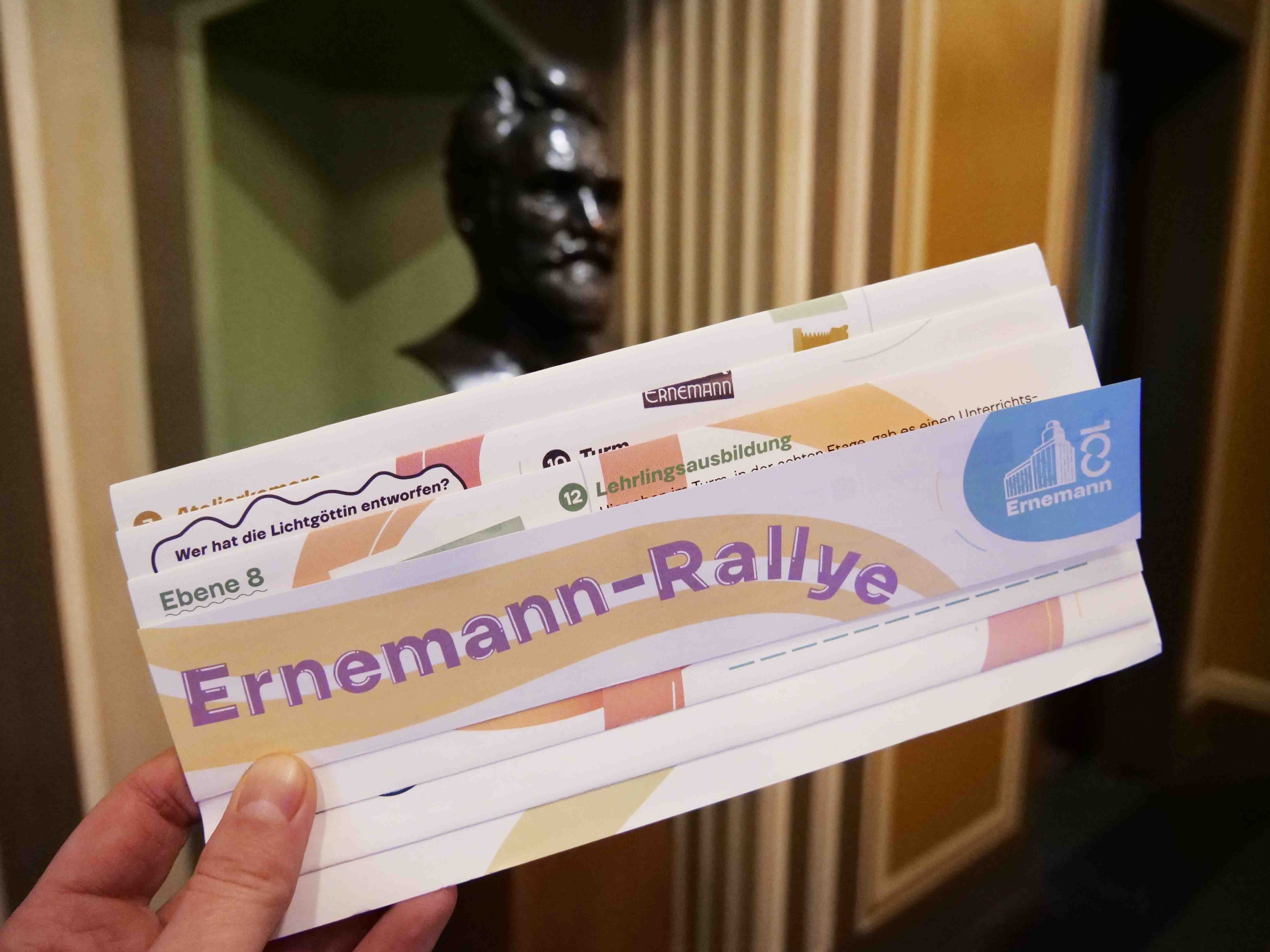 KI generiert: Das Bild zeigt eine Hand, die mehrere Karten mit der Aufschrift "Ernemann-Rallye" hält. Im Hintergrund ist eine metallische Büste zu sehen.