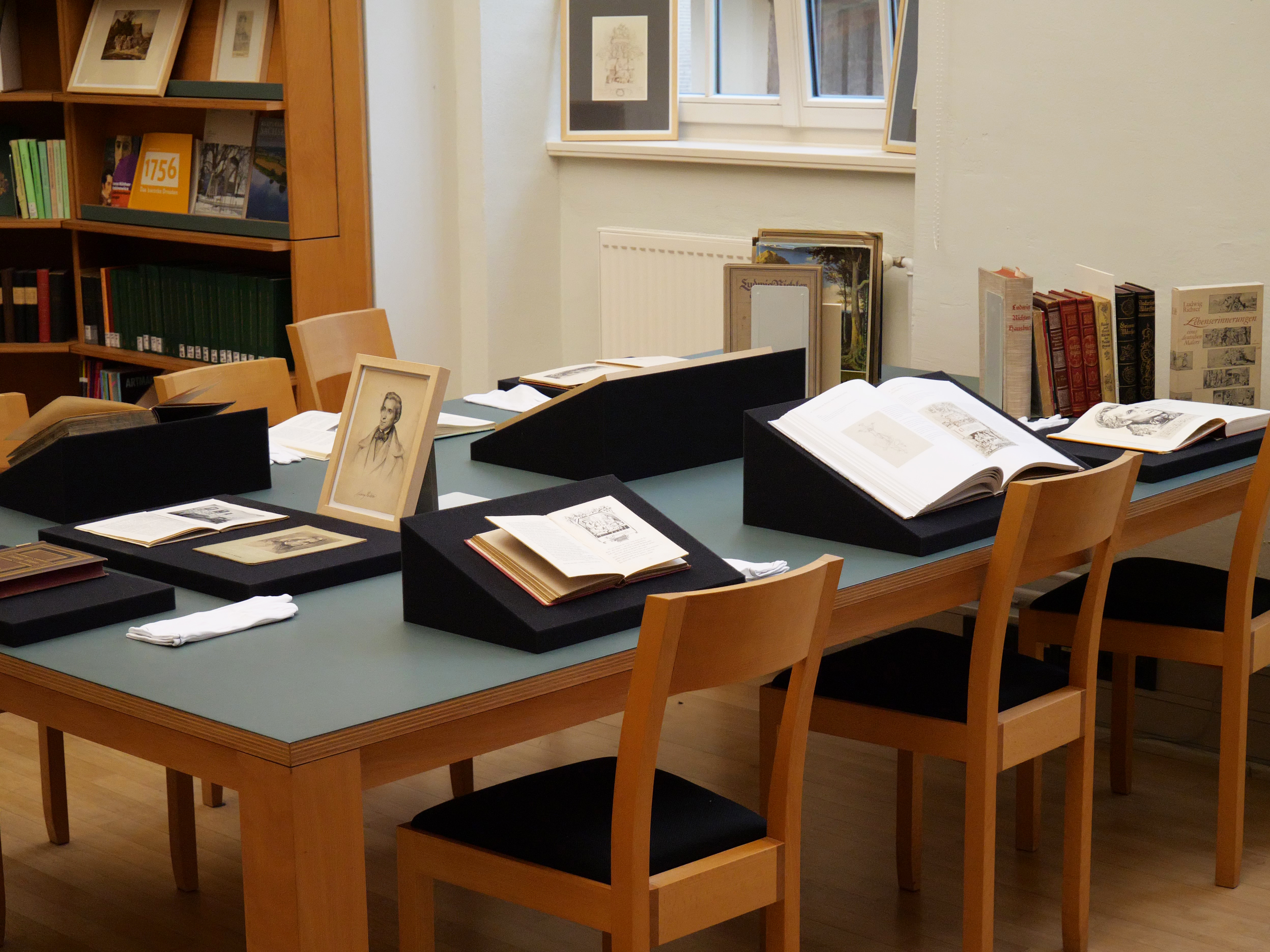Generated by AI: The image shows a study area in a library with several old books open on a table. Framed pictures hang on the walls, and shelves full of books can be seen in the background.