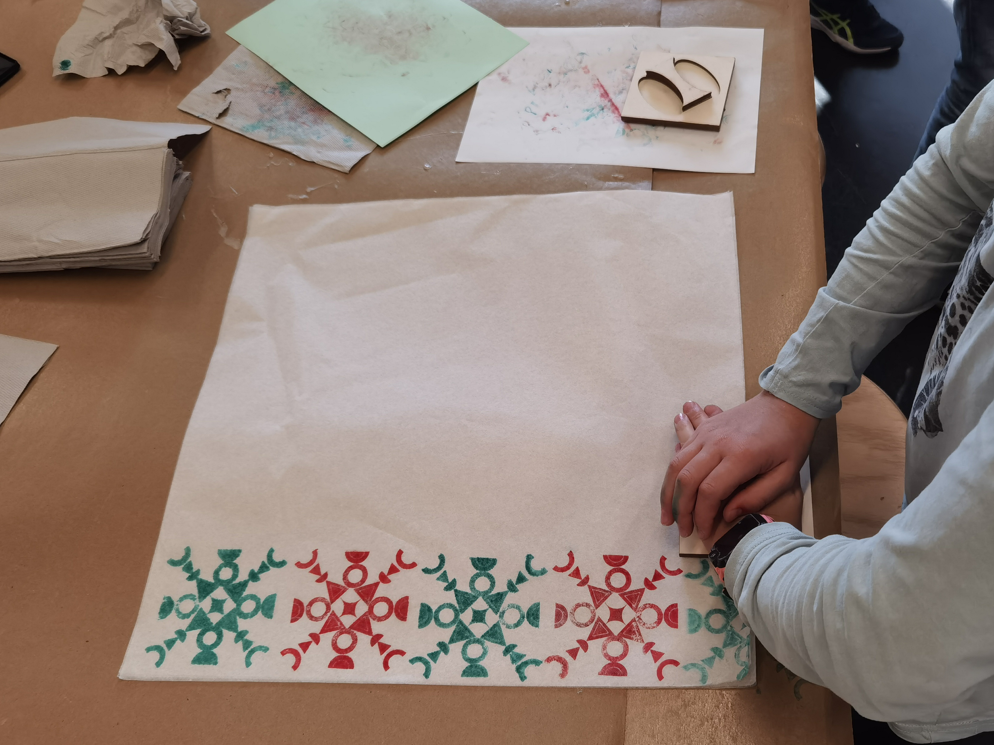 KI generiert: Ein Kind stempelt ein symmetrisches Muster in Rot und Grün auf ein Blatt Papier. Der Fokus liegt auf dem kreativen Prozess und den dekorativen Mustern.