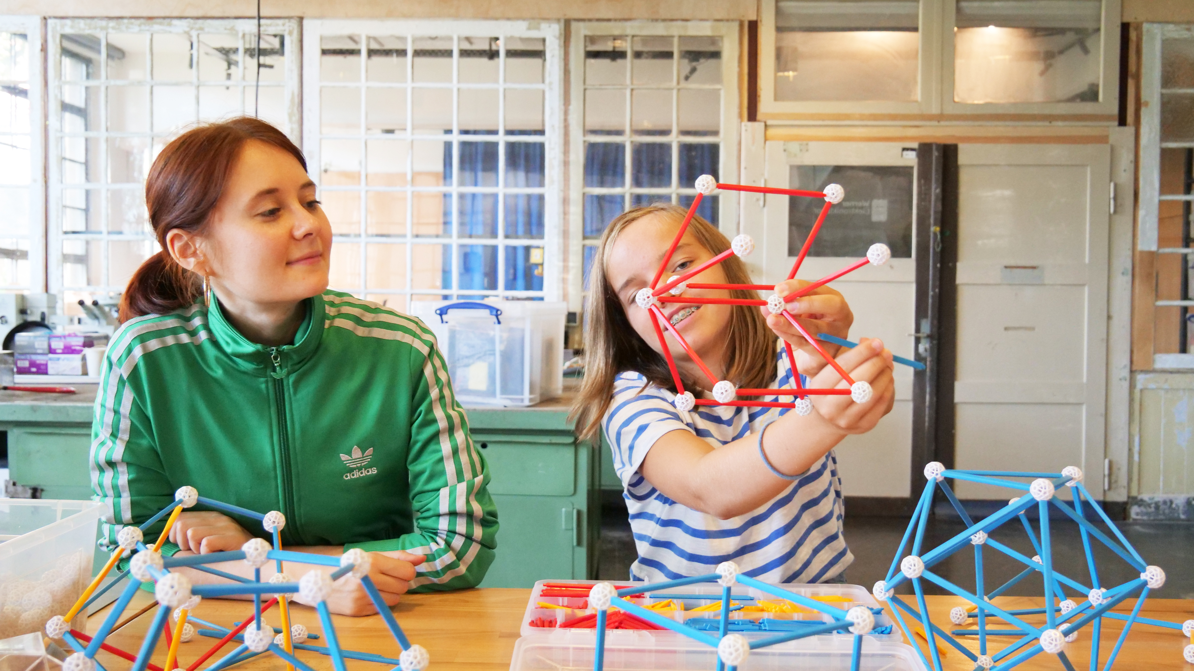 KI generiert: Das Bild zeigt zwei Personen, die mit geometrischen Bausätzen aus Stäben und Verbindungskugeln eine Konstruktion erstellen. Der Fokus liegt auf der Basteltätigkeit und dem gemeinsamen Spaß an der geometrischen Gestaltung.