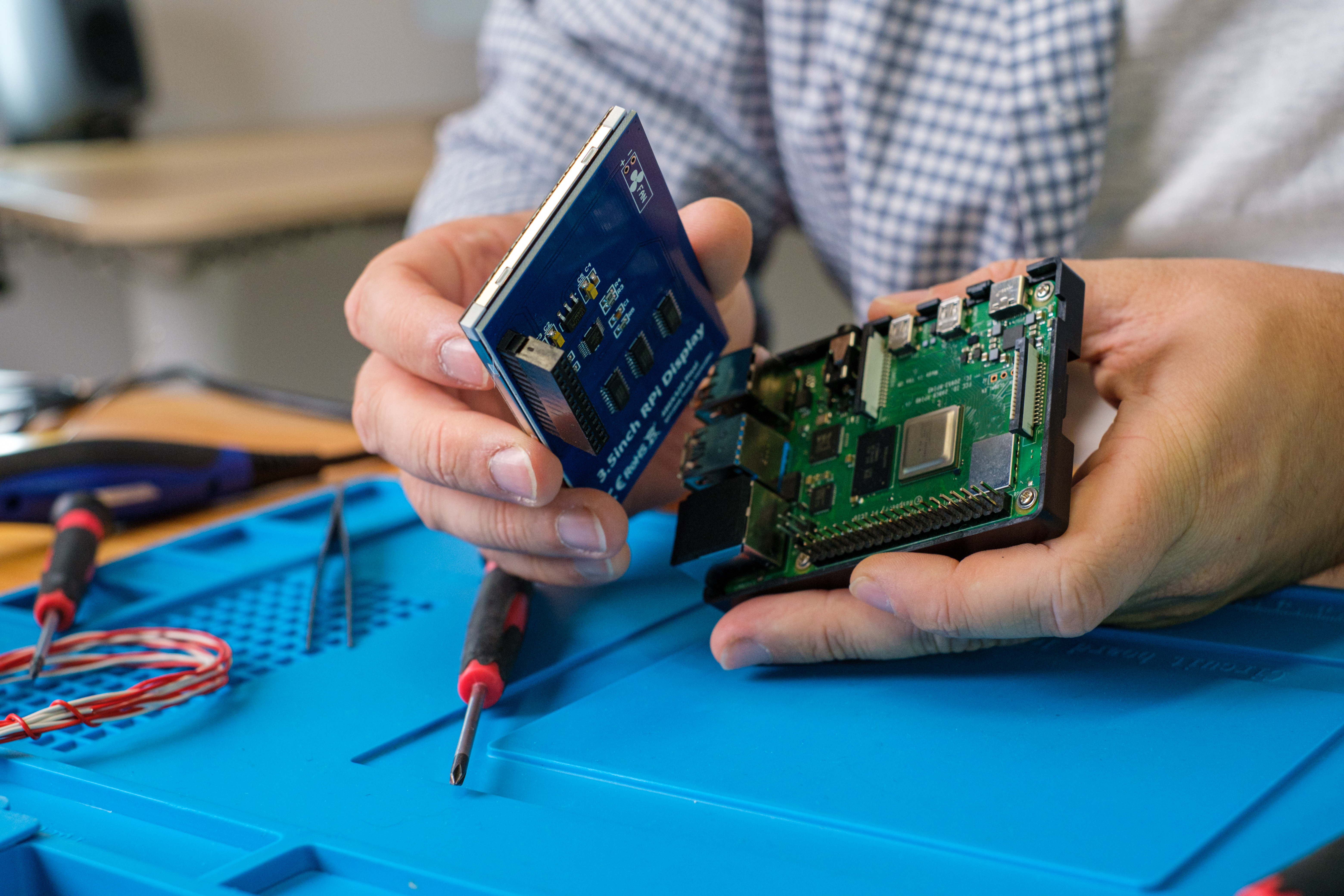 KI generiert: Das Bild zeigt eine Person, die ein elektronisches Gerät, möglicherweise einen Einplatinencomputer, hält und bearbeitet. Im Hintergrund sind Werkzeuge und eine Arbeitsmatte zu sehen, die darauf hindeuten, dass es sich um eine Technik- oder Bastelarbeit handelt.