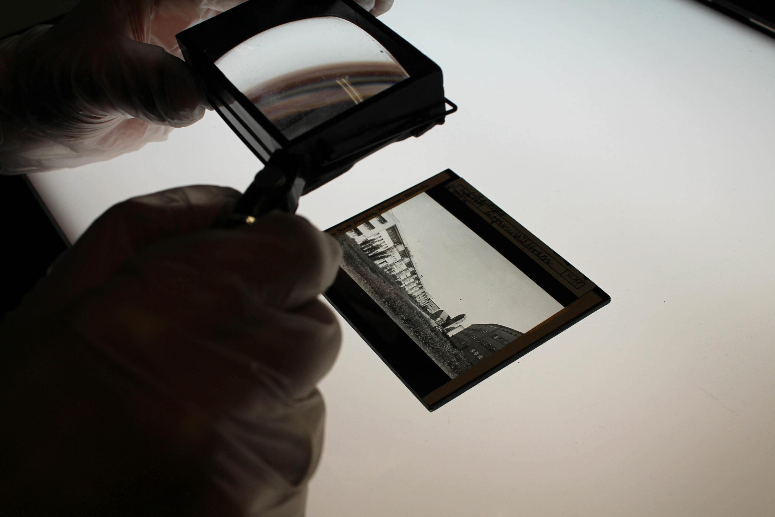 AI generated: The image shows a person wearing gloves examining a photo negative (a black and white glass plate), using a magnifying glass. The scene probably takes place on a light table to make the negative more visible.