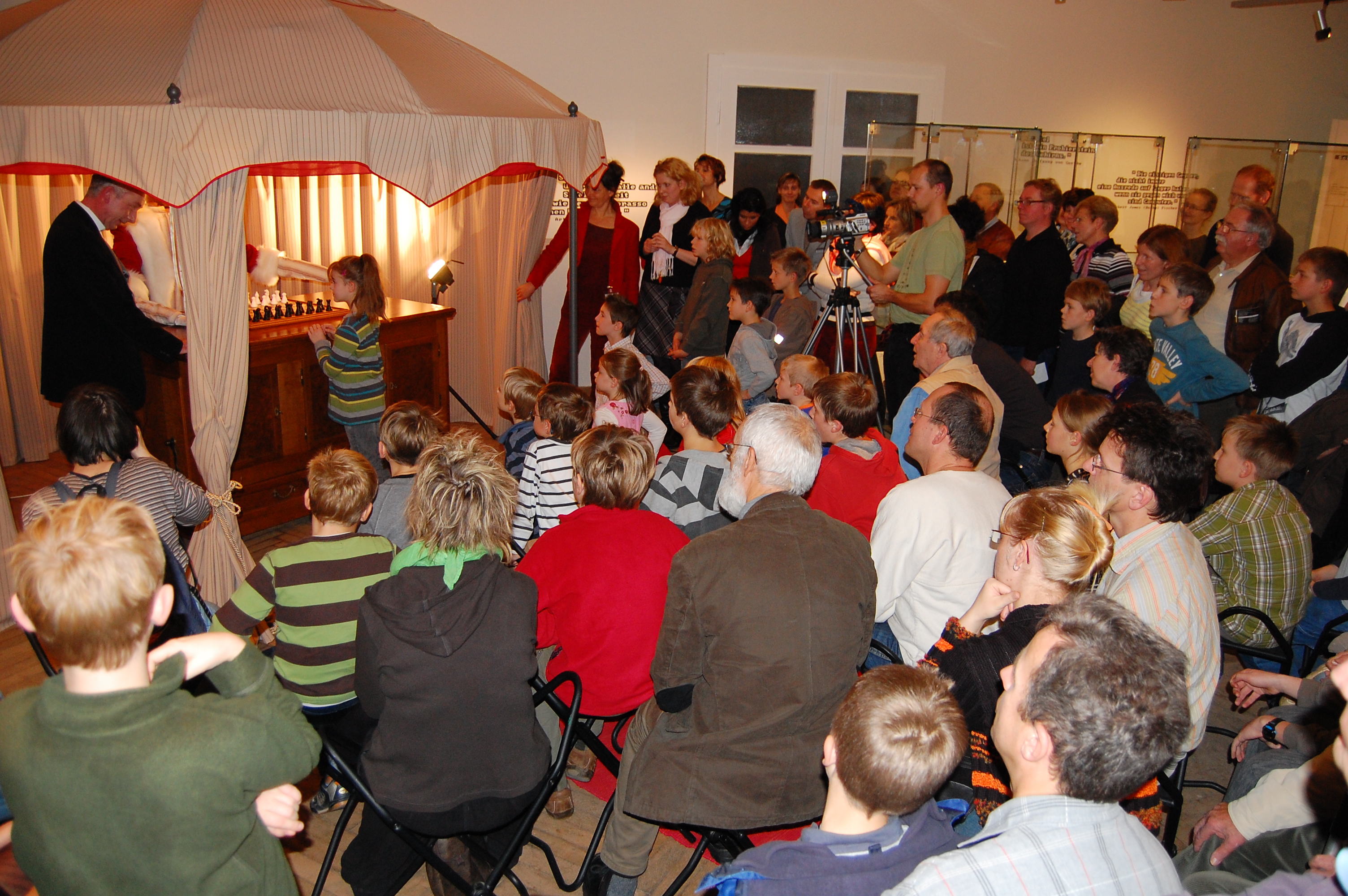 KI generiert: Das Bild zeigt eine Gruppe von Menschen, die einem Mann zuschauen, der sich in einem Pavillon befindet und ein Schachbrett demonstriert. Die Zuschauer sitzen und stehen dicht gedrängt, um die Vorführung zu beobachten.
