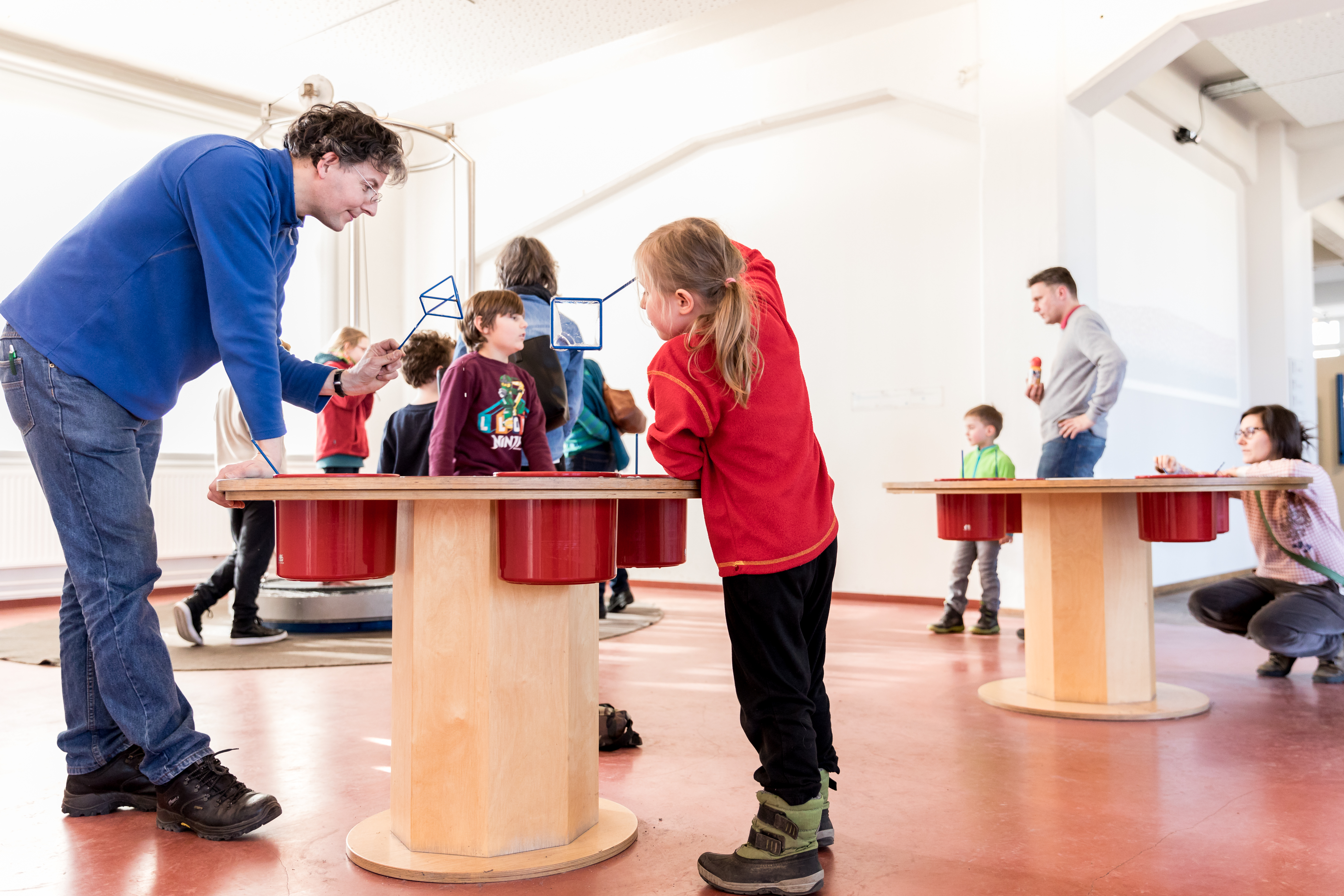 KI generiert: Das Bild zeigt eine Gruppe von Erwachsenen und Kindern, die in einem Raum mit interaktiven Exponaten spielen und lernen. Im Vordergrund interagiert ein Erwachsener mit einem Kind an einem Tisch.