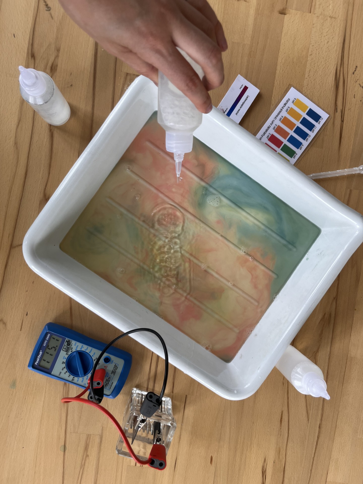 KI generiert: Das Bild zeigt ein wissenschaftliches Experiment, bei dem eine Flüssigkeit in eine Schale mit Wasser und Farbstoffen getropft wird. Es sind ein Multimeter, Elektroden, sowie pH-Teststreifen zu sehen, was auf eine Untersuchung von elektrischen und chemischen Eigenschaften hindeutet.