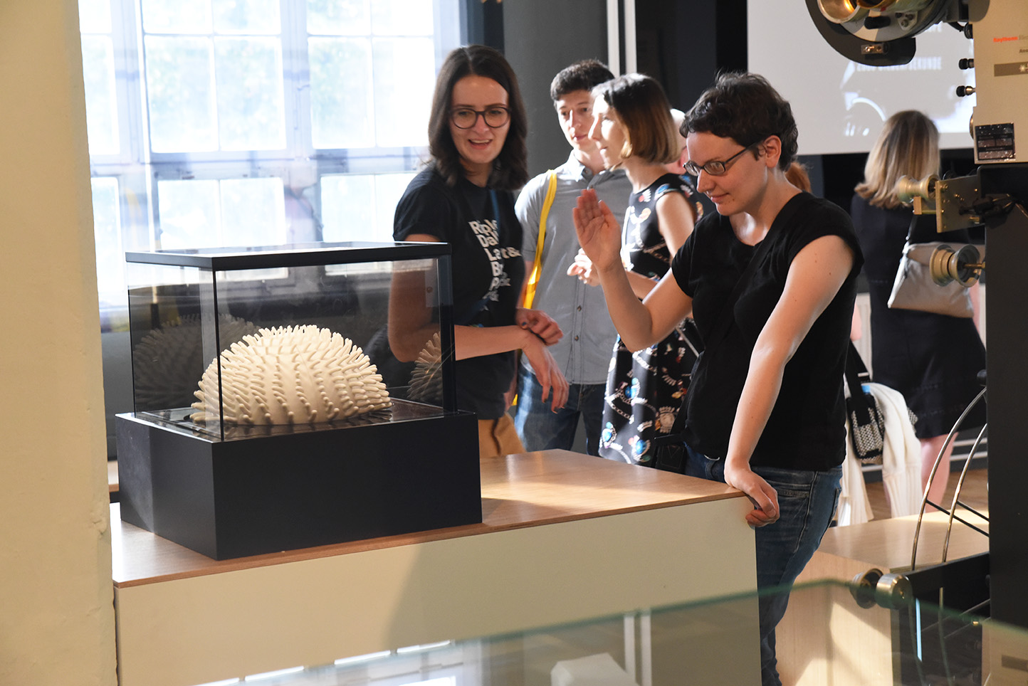 KI generiert: Das Bild zeigt mehrere Personen in einem Ausstellungsraum, die eine Skulptur in einem Glaskasten betrachten. Im Hintergrund sind weitere Exponate und Besucher zu sehen.