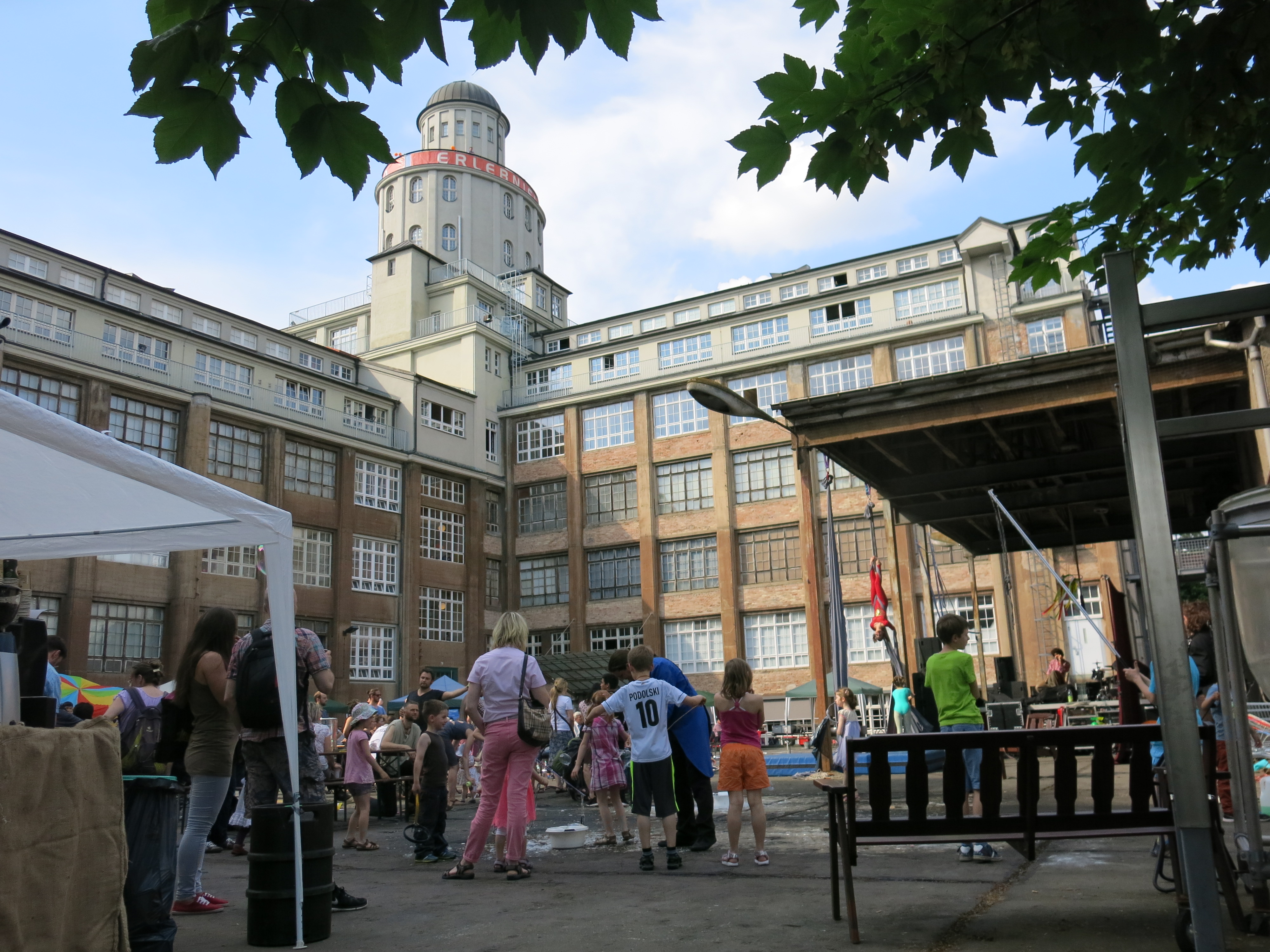 KI generiert: Das Bild zeigt eine Außenansicht eines großen, mehrstöckigen Gebäudes mit einem Turm, umgeben von Bäumen. Im Vordergrund versammeln sich viele Menschen, darunter Kinder, auf einem Platz, scheinbar bei einem Fest oder einer Gemeinschaftsveranstaltung.