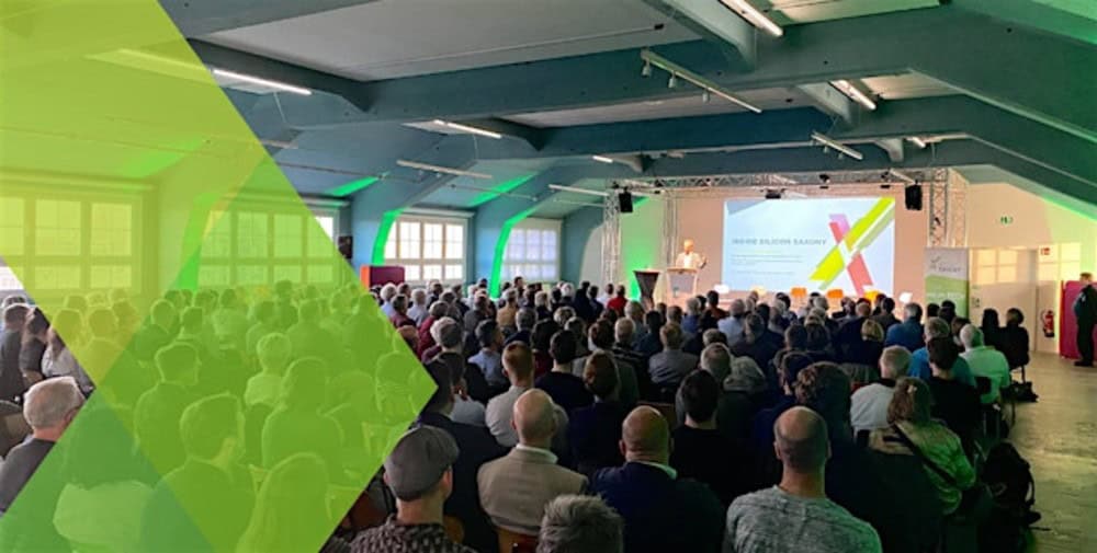 Generated by AI: The image shows a conference or presentation with a speaker on stage and a large audience in a hall. A green graphic element is superimposed over the left-hand side of the image.