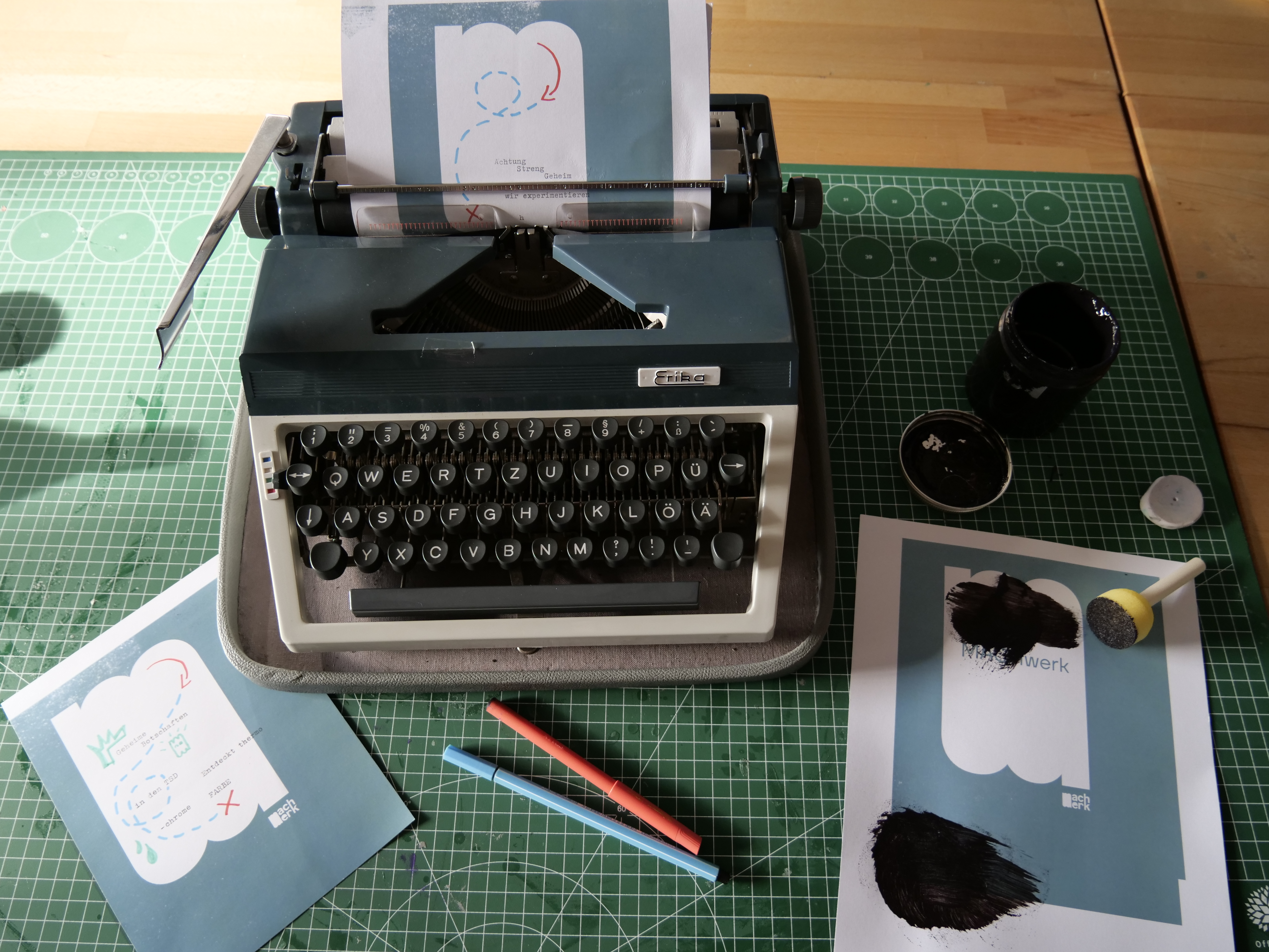 KI generiert: Das Bild zeigt eine alte Schreibmaschine, auf der ein Blatt Papier eingespannt ist, umgeben von Zeichenutensilien und Zeichnungen auf einem Schneidemattentisch. Es vermittelt eine kreative Arbeitsatmosphäre mit einem Mix aus analogem Schreiben und Malen.