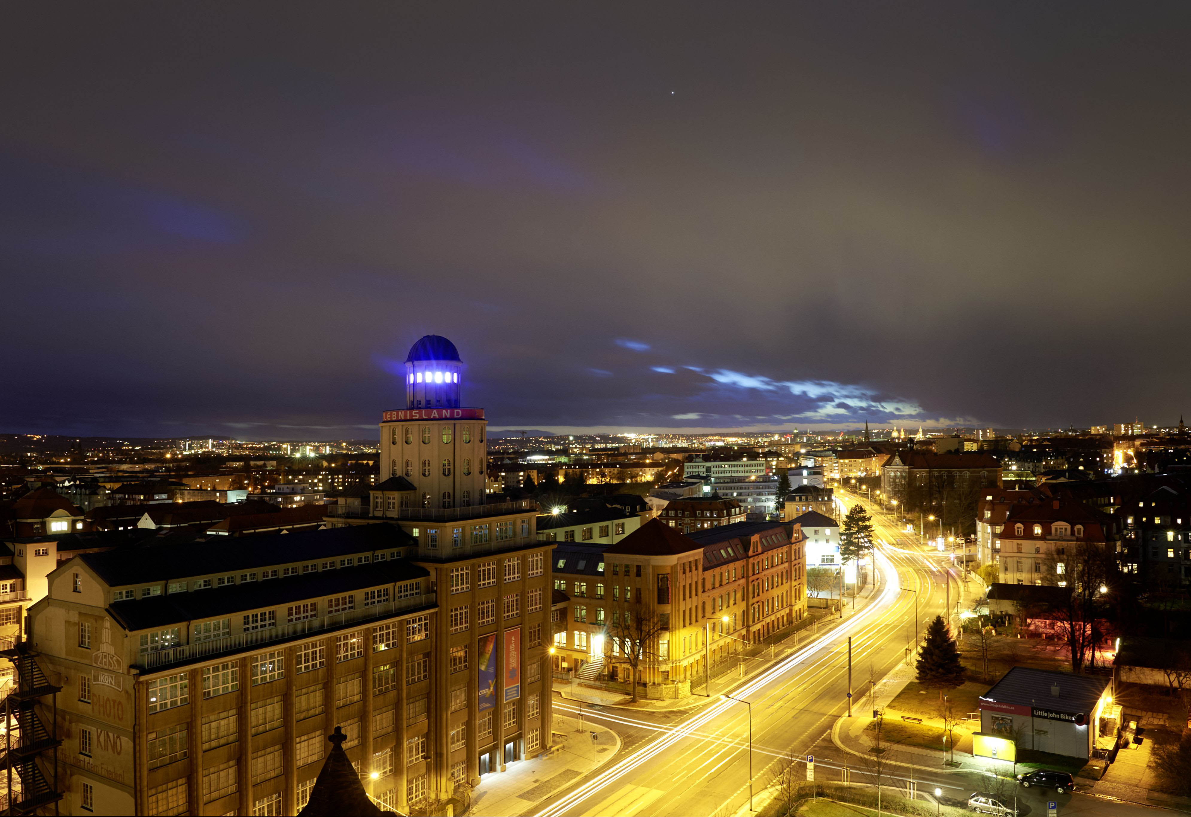 KI generiert: Das Bild zeigt eine beleuchtete Stadtansicht bei Nacht, im Vordergrund ein markantes Gebäude mit einem Turm, auf dem "MIELISLAND" geschrieben steht. Der Verkehr hinterlässt Lichtspuren auf den Straßen, und am Horizont sind weitere Lichter der Stadt sichtbar.