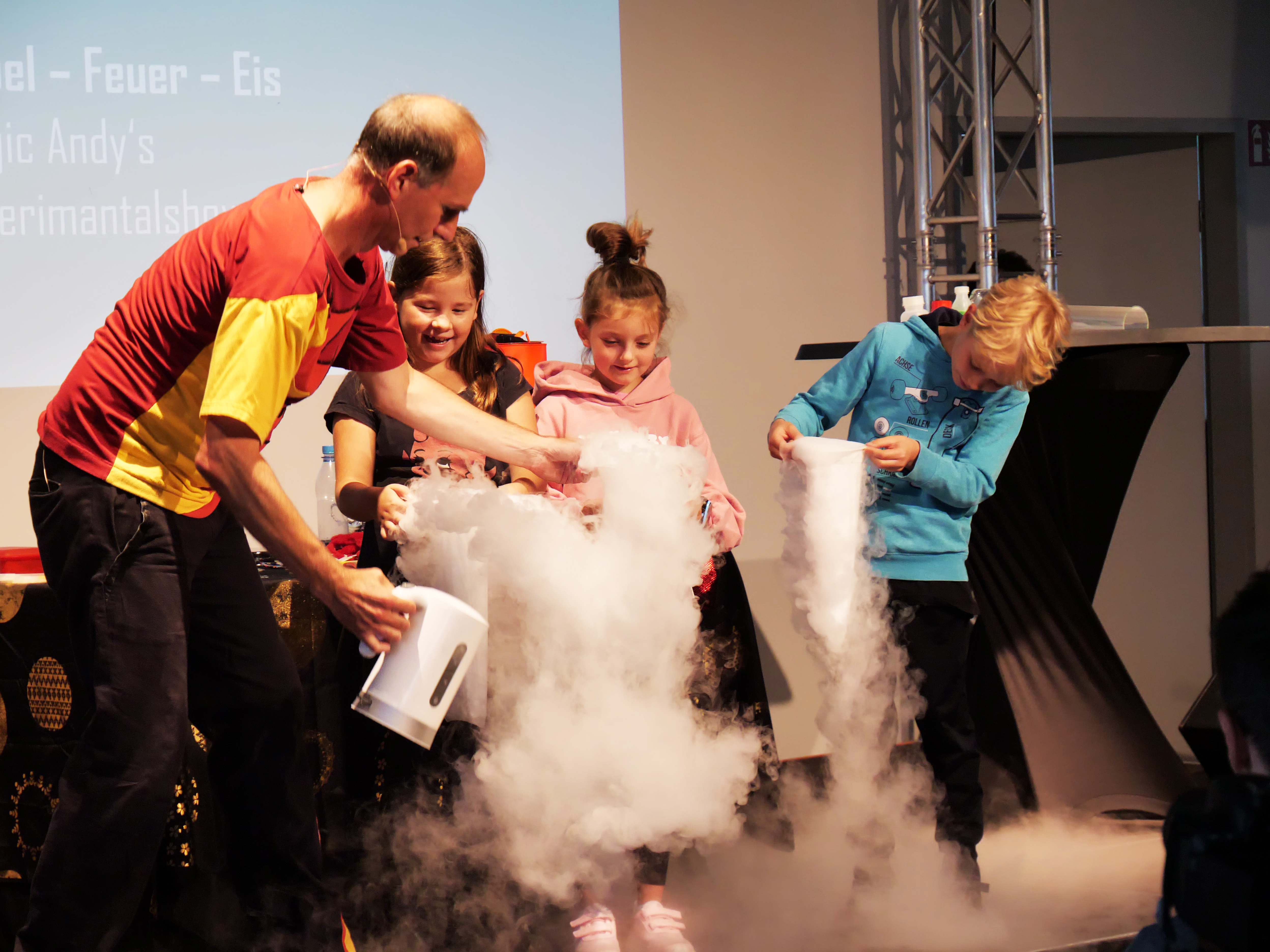 KI generiert: Das Bild zeigt eine Experimentiershow, bei der ein Erwachsener mit drei Kindern ein wissenschaftliches Experiment durchführt, in dem dicker Nebel entsteht. Die Kinder scheinen fasziniert von dem Effekt zu sein.