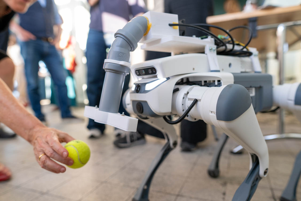 KI generiert: Das Bild zeigt einen Roboterhund mit einem mechanischen Arm, der einer Person gegenübersteht, die ihm einen gelben Tennisball hinhält. Im Hintergrund sind unscharf stehende Personen zu sehen.