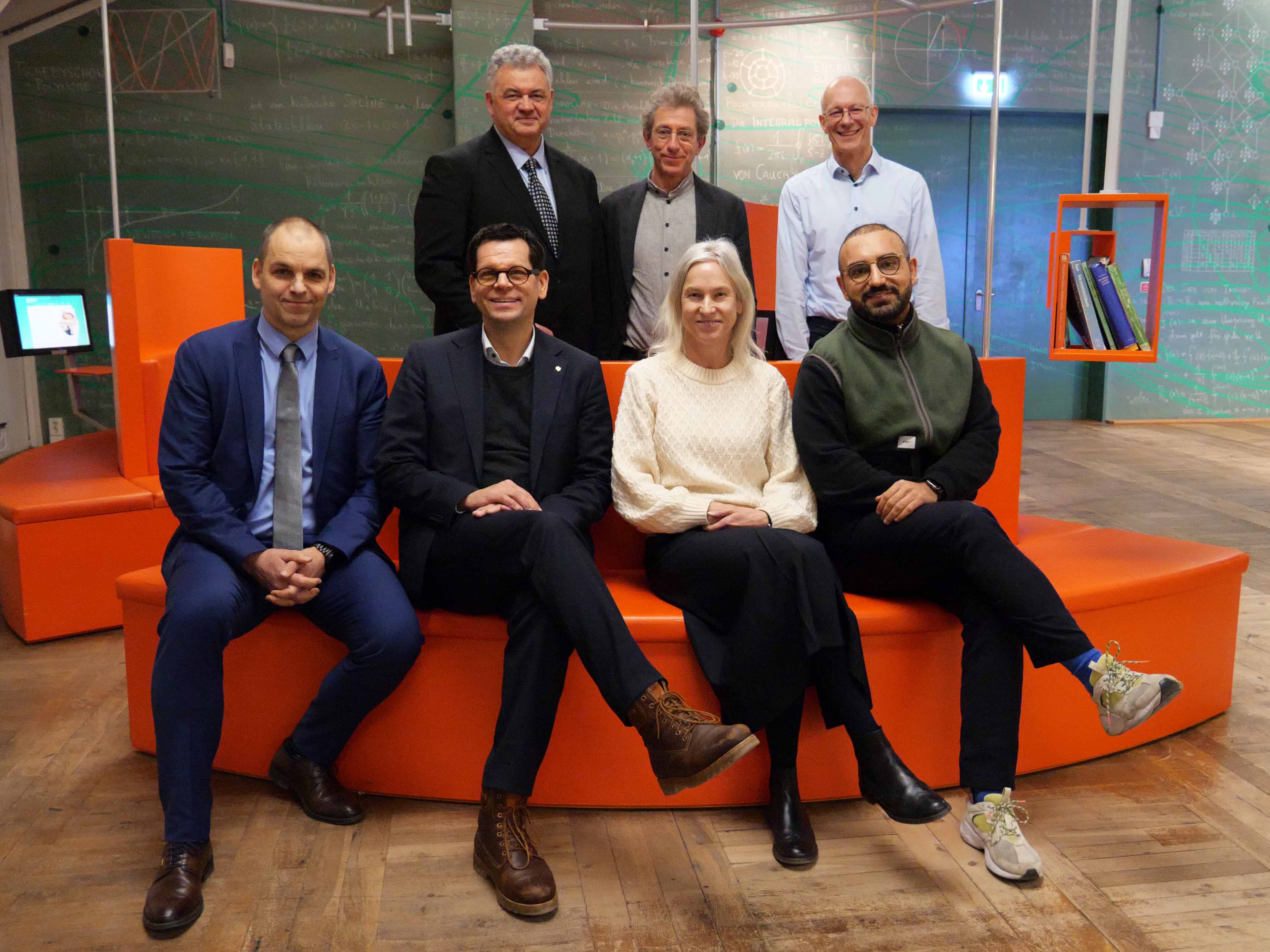 AI generated: The image shows seven people standing and sitting in a modern room with orange-coloured seating and writable walls. They pose together for a group photo.