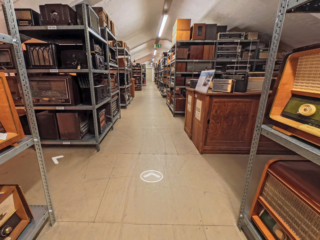 KI generiert: Das Bild zeigt einen Raum voller Regale mit alten Radios und elektronischen Geräten. Der Gang ist beleuchtet und die Regale sind dicht mit diesen Sammlerstücken gefüllt.