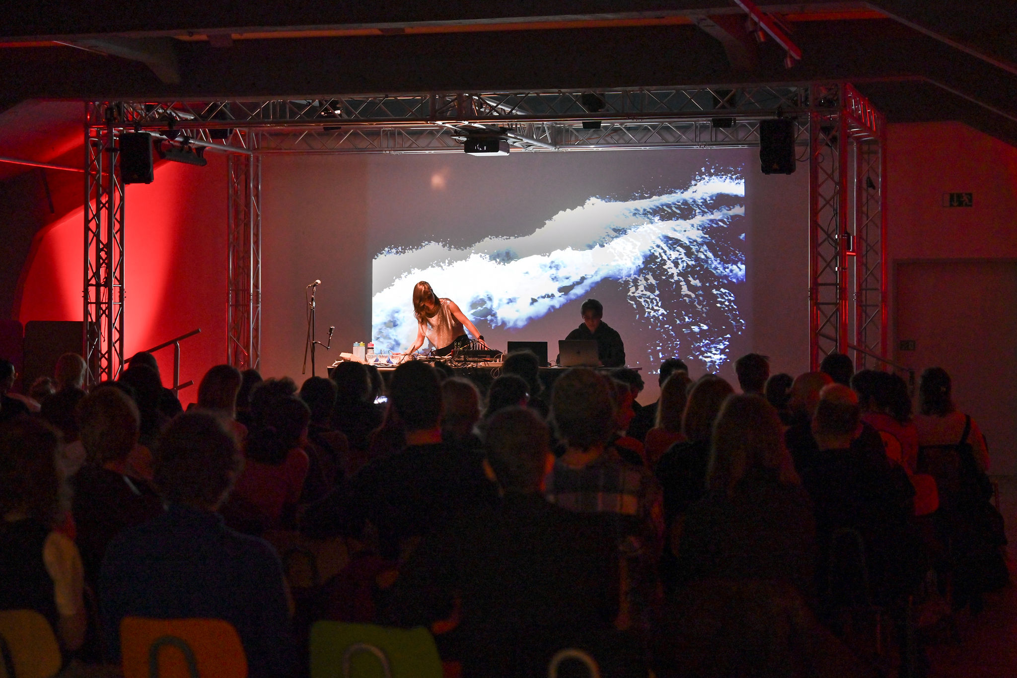 KI generiert: Das Bild zeigt eine Live-Performance mit zwei Personen auf einer Bühne, während eine Hintergrundprojektion von Meereswellen zu sehen ist. Das Publikum sitzt und schaut aufmerksam zu.