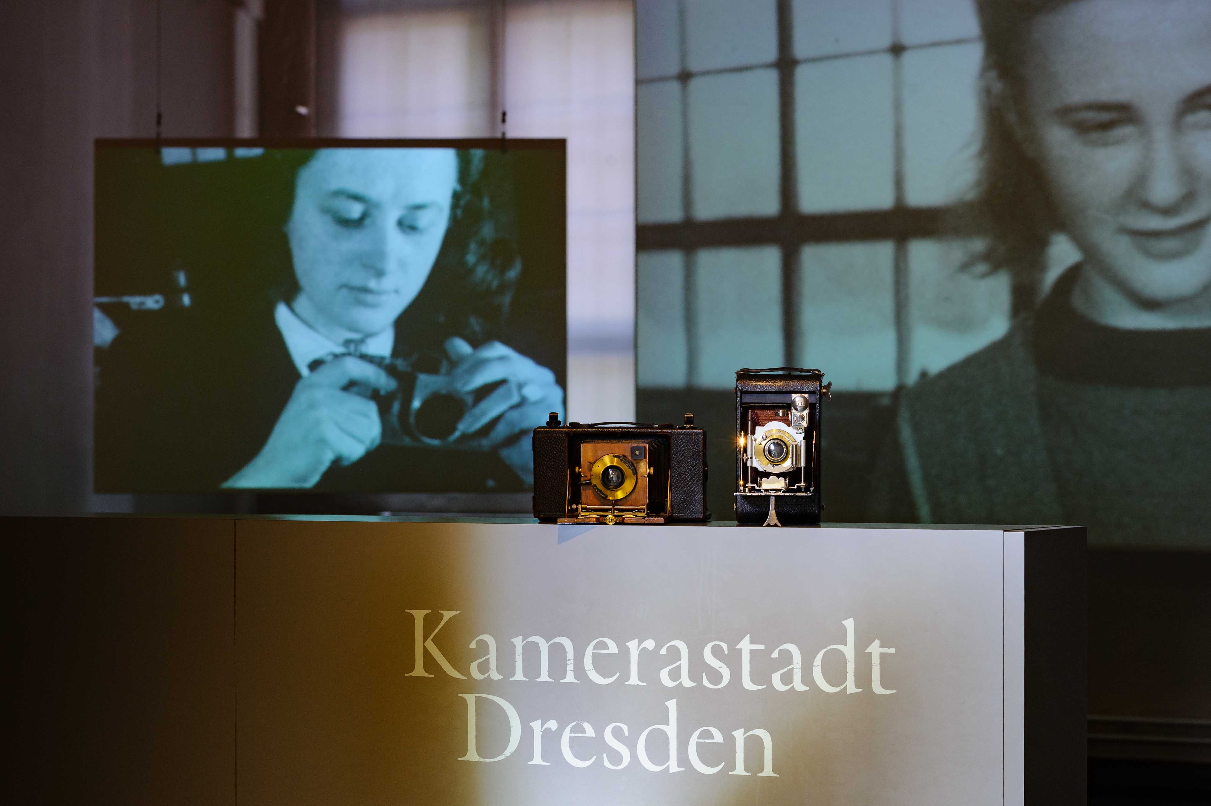 KI generiert: Das Bild zeigt eine Ausstellung mit historischen Kameras und der Beschriftung "Kamerastadt Dresden", wobei im Hintergrund ein Schwarzweißfoto einer Person zu sehen ist, die eine Kamera hält. Dies deutet auf die historische Bedeutung Dresdens in der Kameraherstellerindustrie hin.