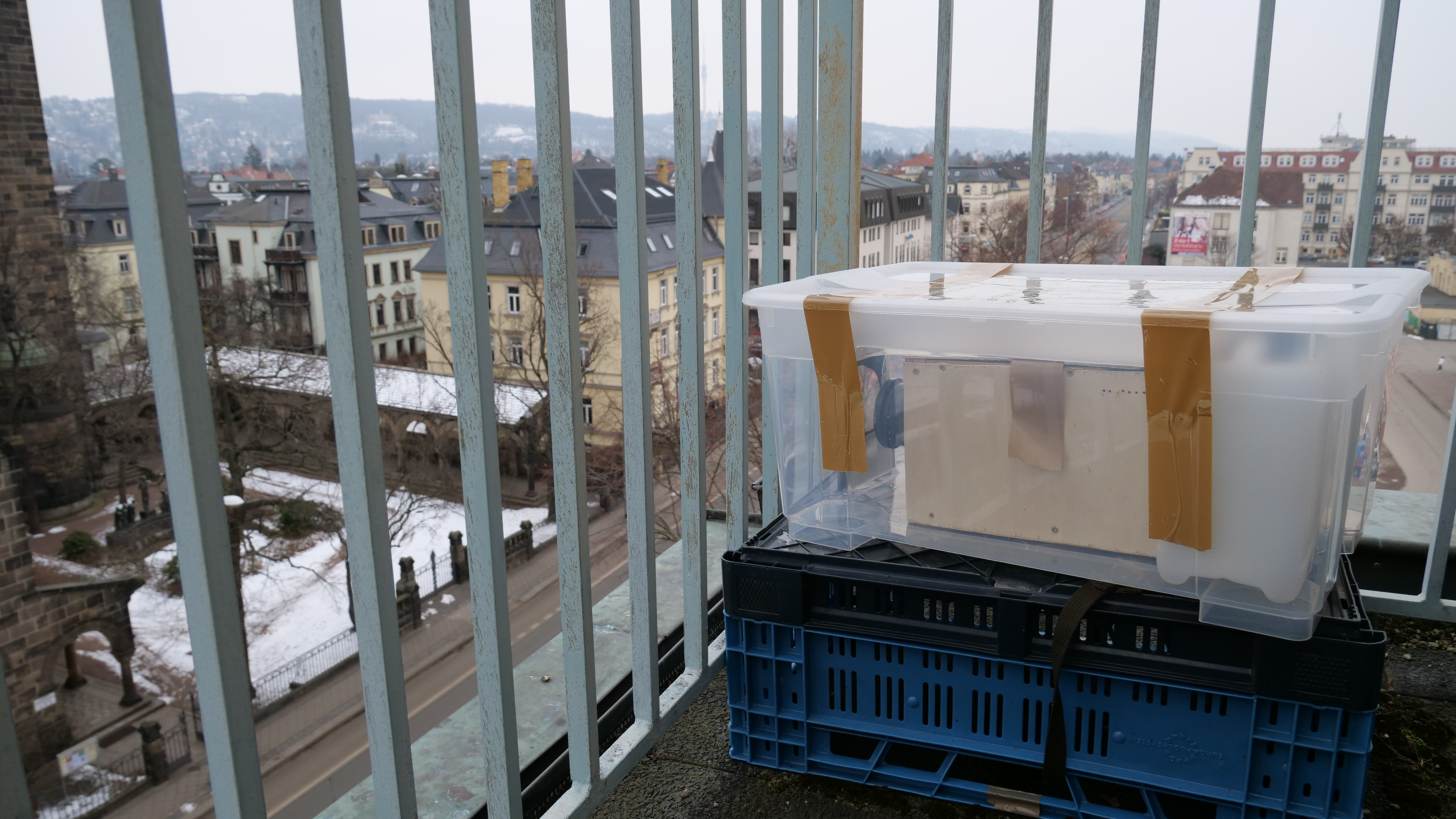 AI generated: The image shows a makeshift construction made of a plastic box on a balcony, held up with adhesive tape and supported by a blue plastic pallet. In the background you can see an urban landscape with several buildings and a slightly snow-covered ground.