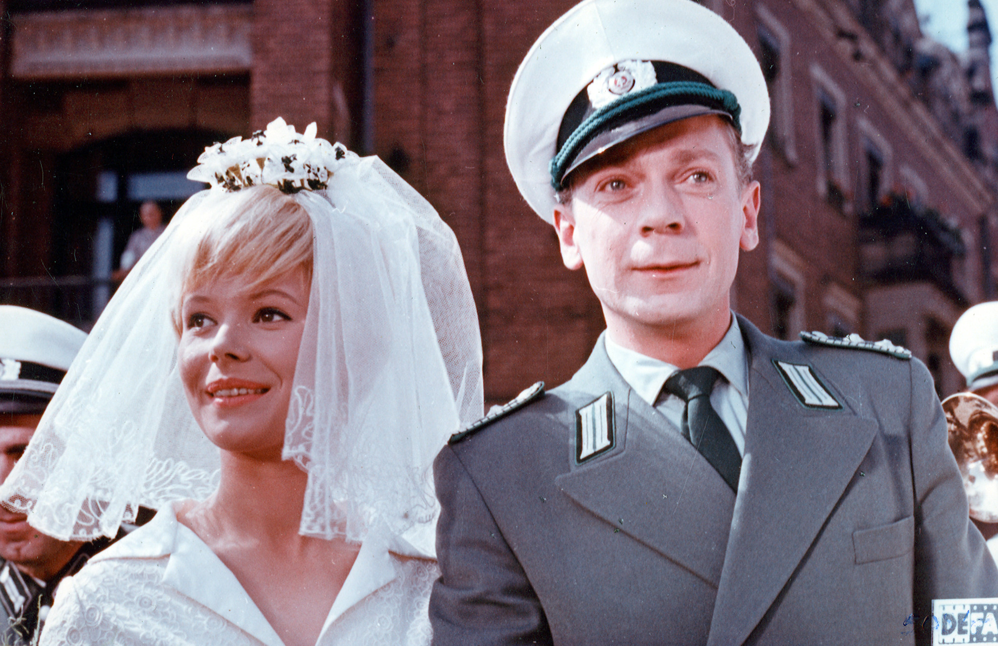 KI generiert: Das Bild zeigt ein Hochzeitspaar, wobei die Frau ein Brautkleid mit Schleier trägt und der Mann eine Uniform. Im Hintergrund sind weitere Personen und Gebäude zu sehen.