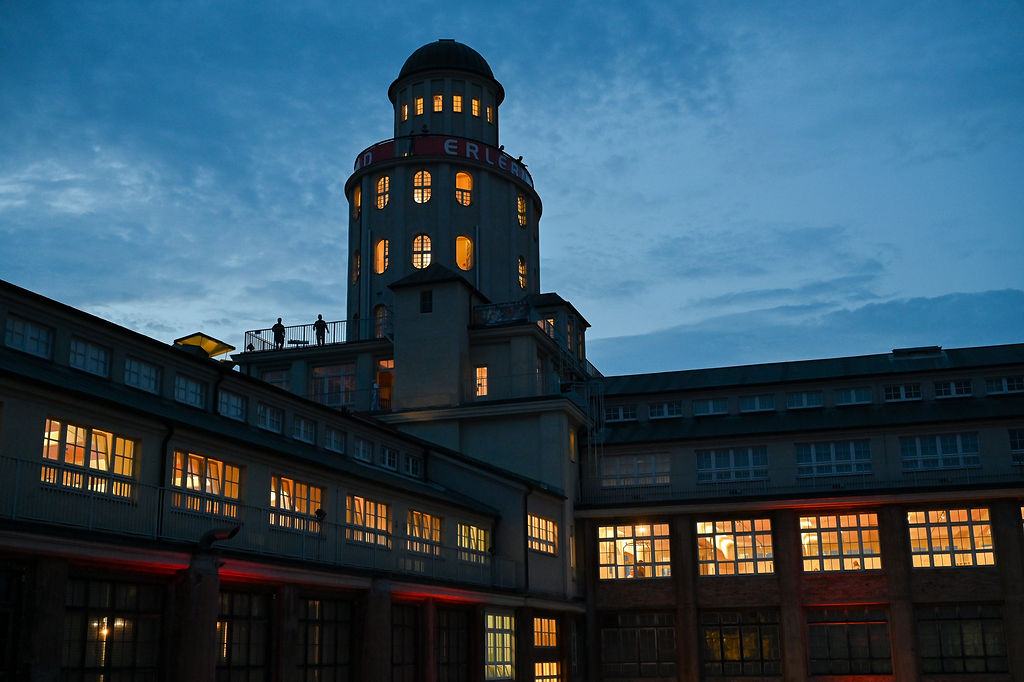 AI generated: The image shows a historic building at dusk with a striking tower and illuminated windows. The sky is slightly cloudy and conveys a calm evening atmosphere.