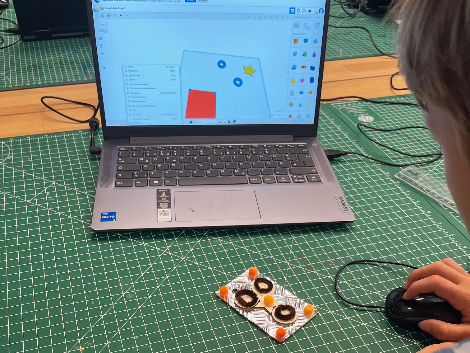 KI generiert: Ein Kind arbeitet an einem Laptop mit einer 3D-Design-Software, während es neben sich ein kleines, auf einer Aluminiumfolie platziertes Bastelprojekt hat. Der Hauptinhalt des Bildes ist das kreative Arbeiten am Computer und das reale Bastelobjekt.