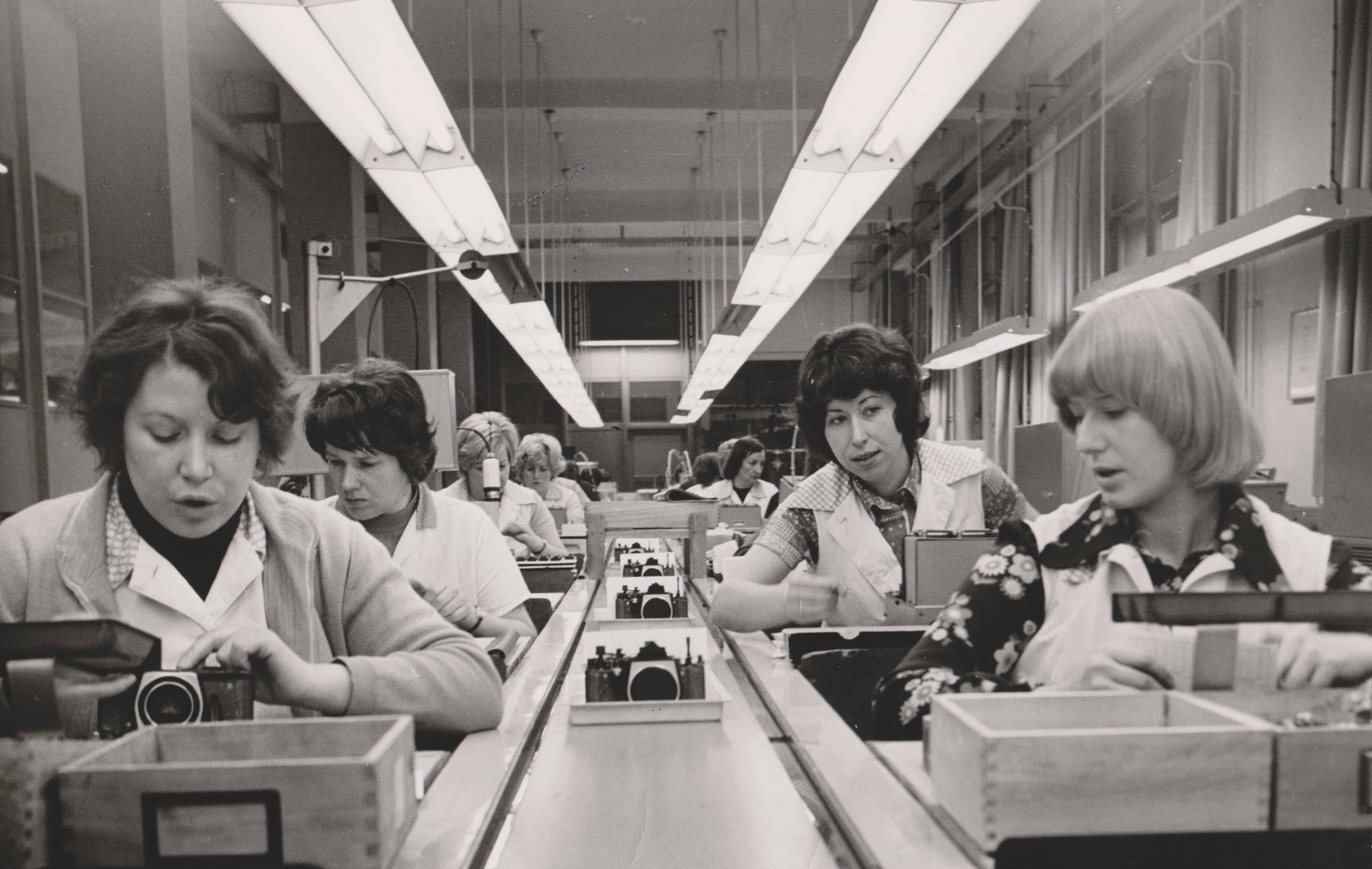 KI generiert: Das Bild zeigt mehrere Frauen in einer Fabrik, die an einer Produktionslinie arbeiten und Kameras zusammenbauen oder überprüfen. Das Hauptthema des Bildes ist die industrielle Fertigung und die Rolle der Arbeiterinnen in diesem Prozess.