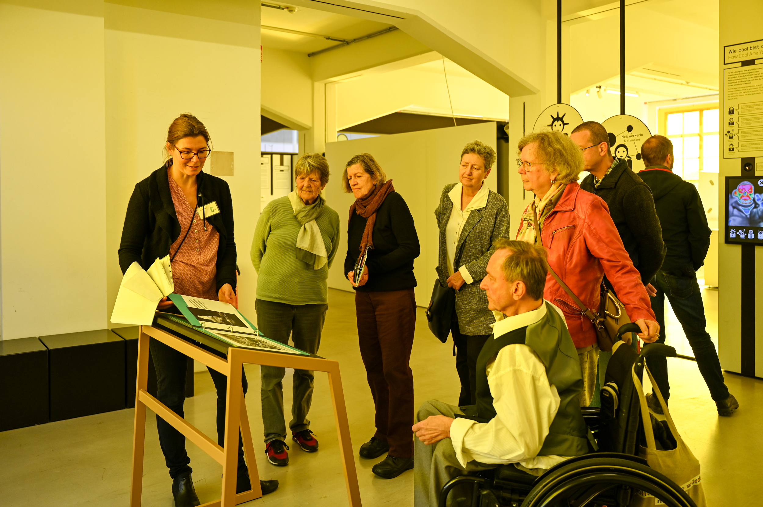 KI generiert: Eine Gruppe von Menschen versammelt sich um einen Ausstellungstisch, während eine Frau etwas präsentiert oder erklärt. Die Umgebung scheint eine Galerie oder ein Museum zu sein.