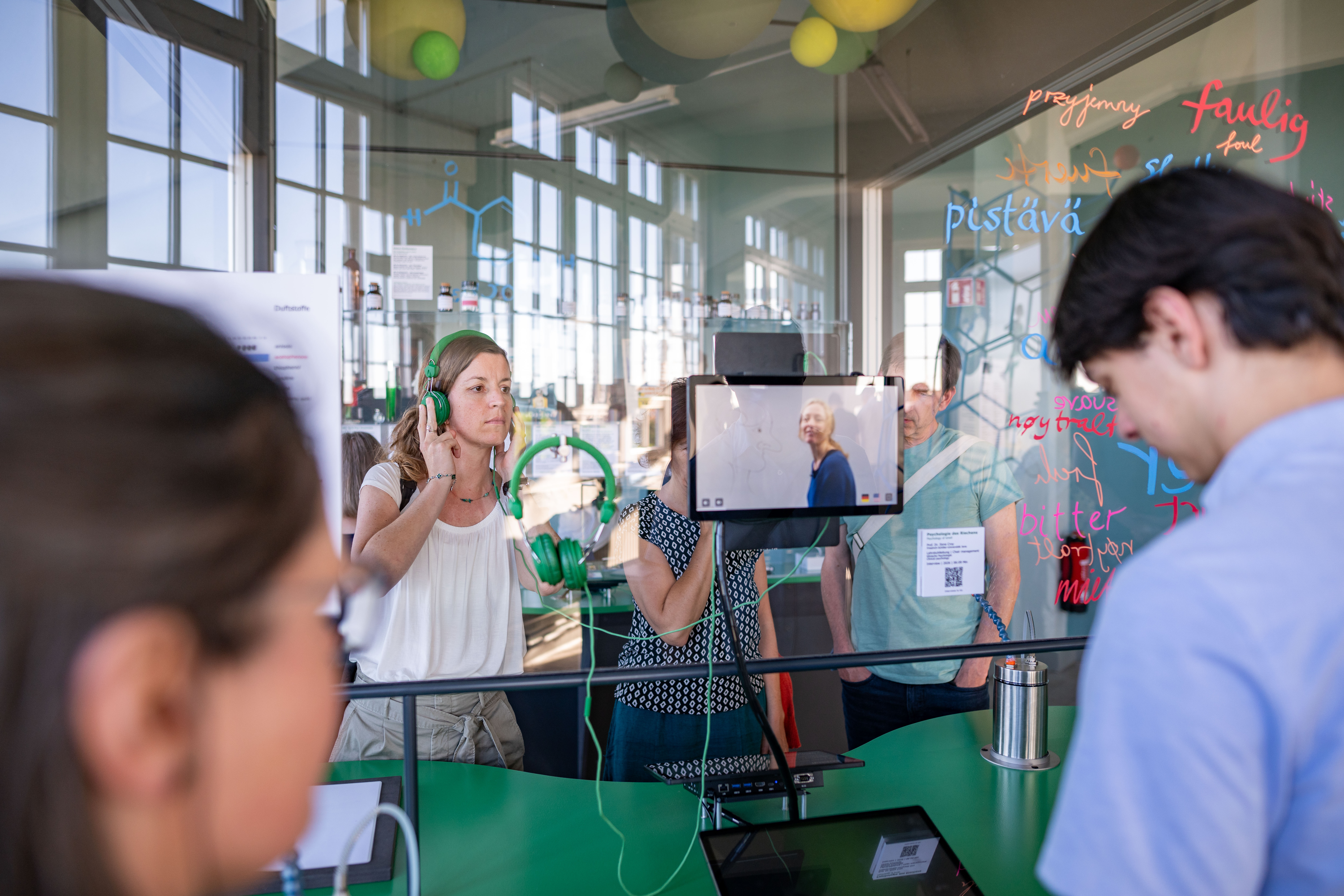 KI generiert: Das Bild zeigt eine Gruppe von Menschen in einer interaktiven Ausstellung, in der sie Geräte nutzen und Informationen auf einem Bildschirm betrachten. Im Hintergrund sind wissenschaftliche Formeln und Begriffe auf einer Glaswand zu sehen.