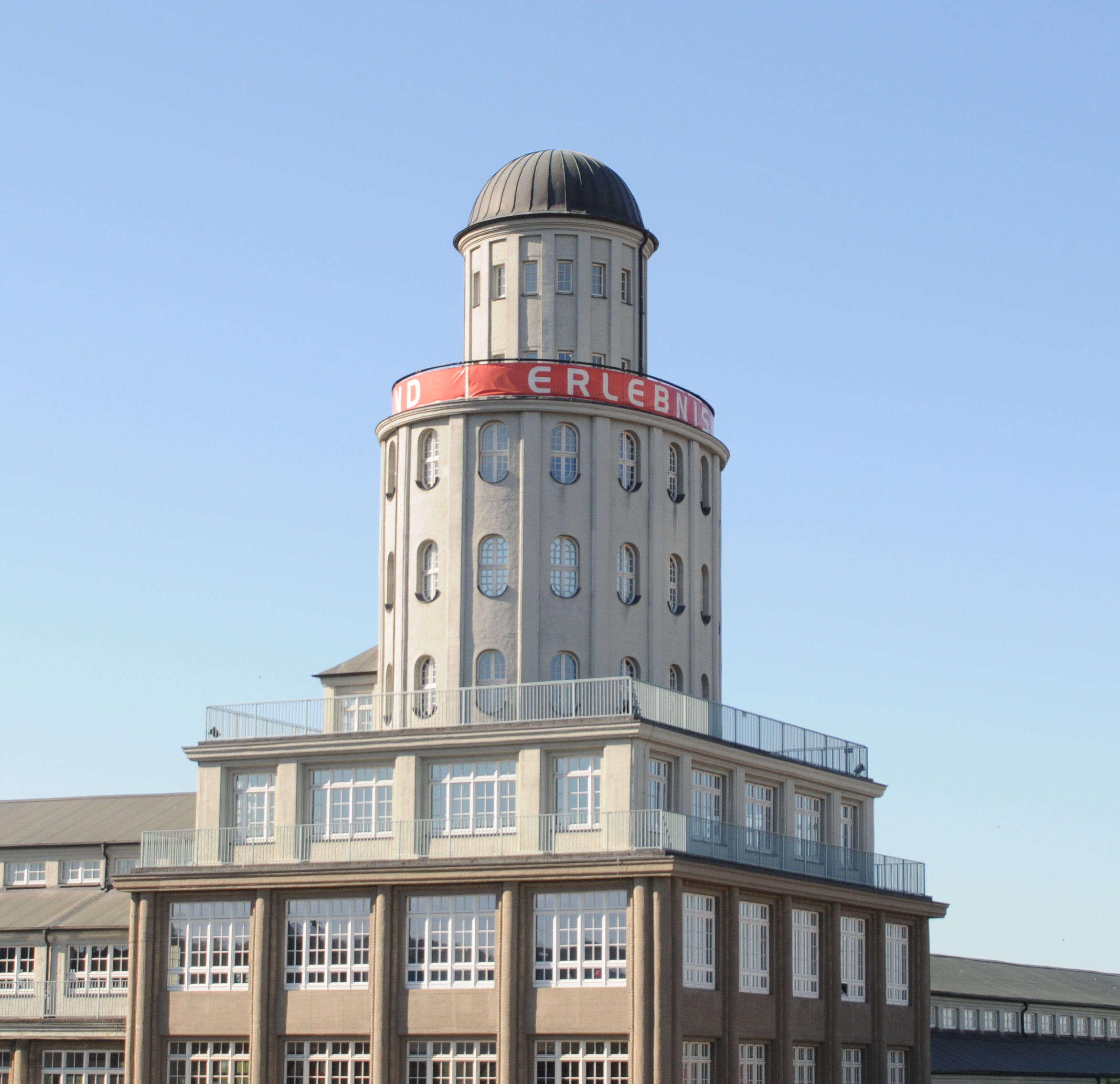 KI generiert: Das Bild zeigt ein mehrstöckiges Gebäude mit einem markanten runden Turm und einer Kuppel auf dem Dach. Der Turm trägt ein rotes Banner mit dem Wort "ERLEBNIS".