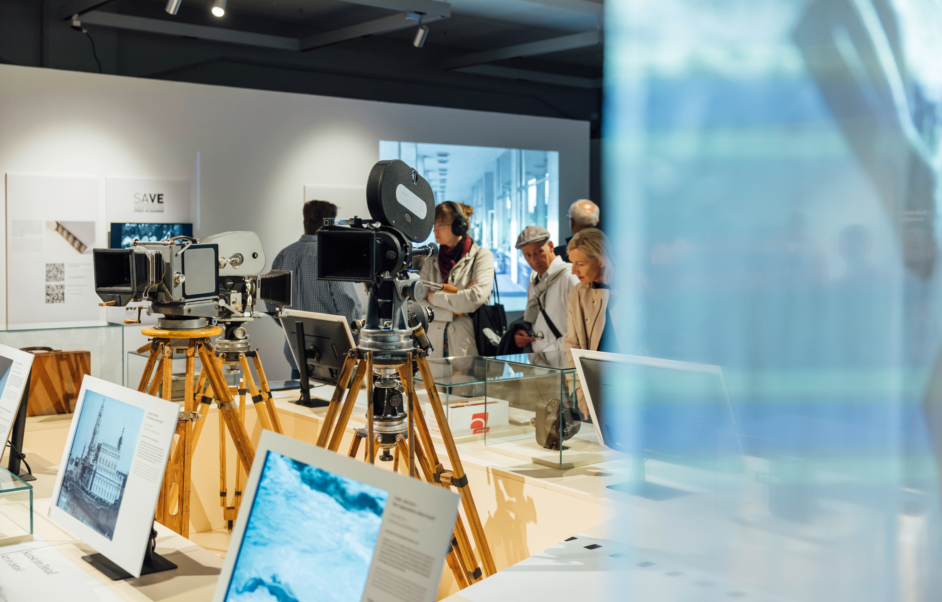 KI generiert: Das Bild zeigt eine Ausstellung von historischen Kameras auf Stativ, die in einem gut beleuchteten Raum präsentiert werden. Im Hintergrund betrachten mehrere Personen interessiert die Exponate.