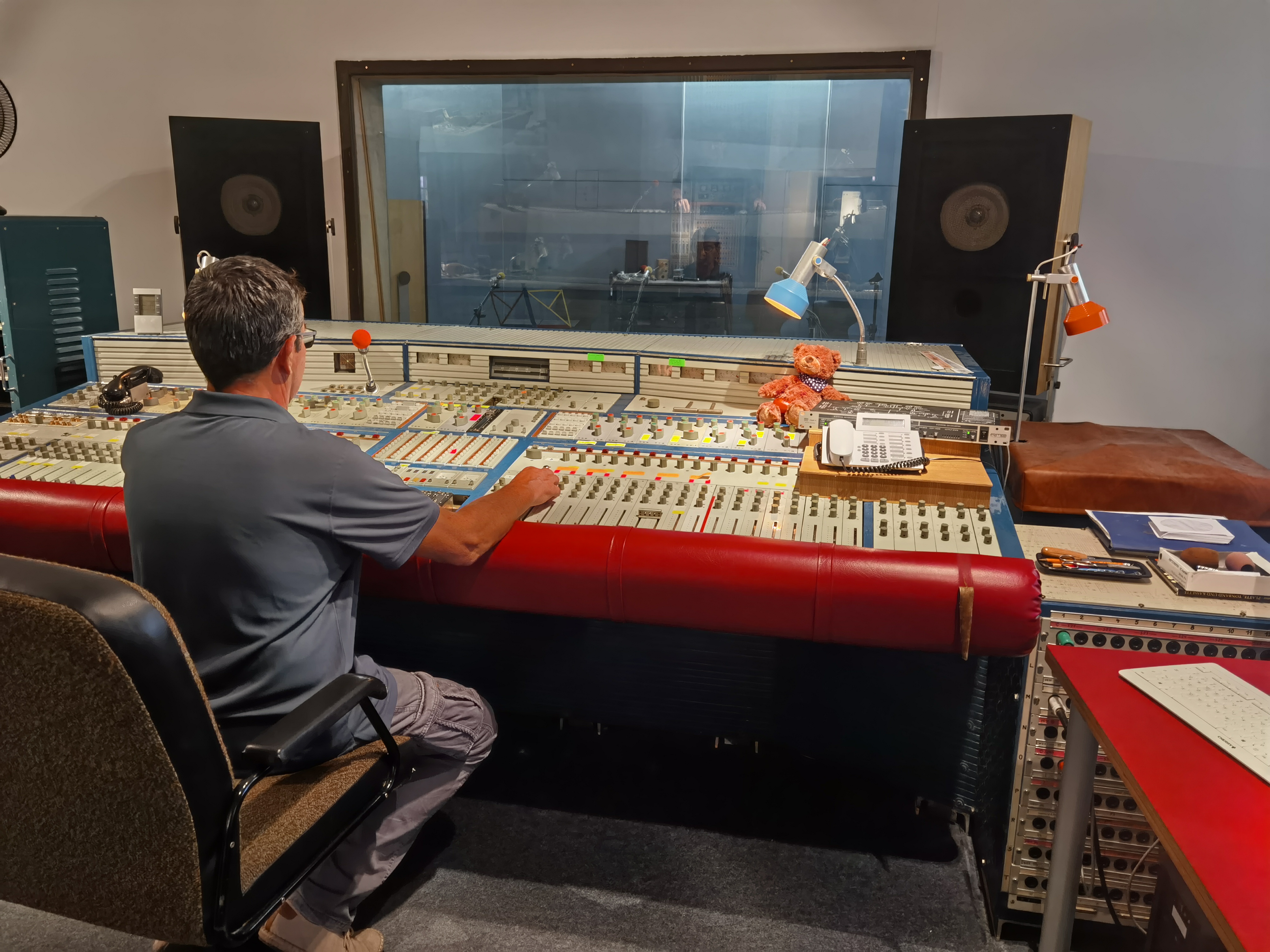 KI generiert: Das Bild zeigt eine Person, die an einem großen Mischpult in einem Tonstudio arbeitet. Im Vordergrund sind zahlreiche Regler und Knöpfe zur Audiosteuerung sichtbar.