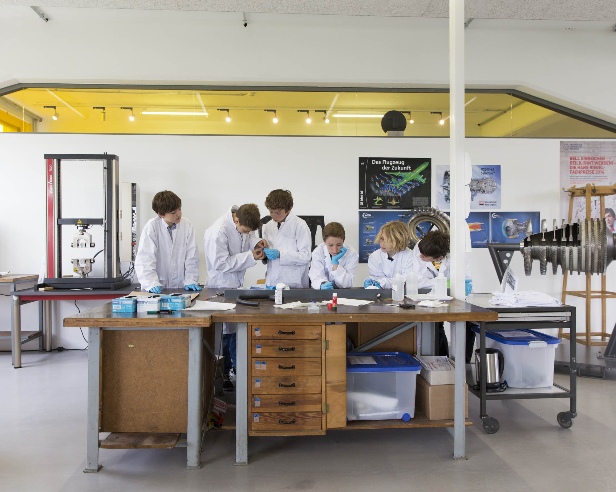 KI generiert: Das Bild zeigt eine Gruppe von Menschen in weißen Laborkitteln, die in einem Labor arbeiten und etwas auf einem Tisch untersuchen. Der Hintergrund deutet auf ein wissenschaftliches Umfeld mit Postern und technischen Ausrüstungen hin.